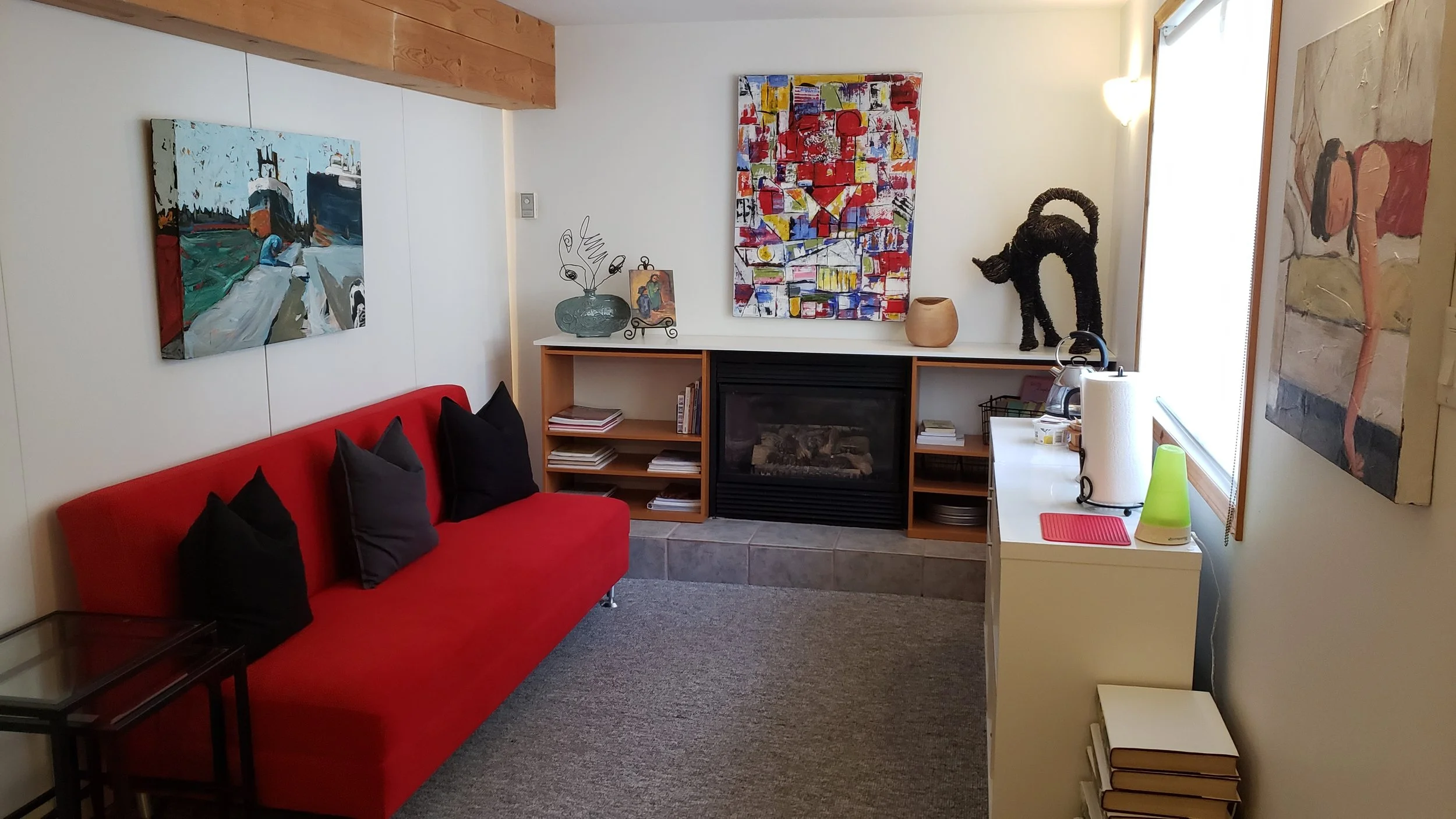 A living room with a red couch, black pillows, abstract paintings on the wall, a fireplace, and decorative items on a shelf, including a sculpture of a black dog and vases.