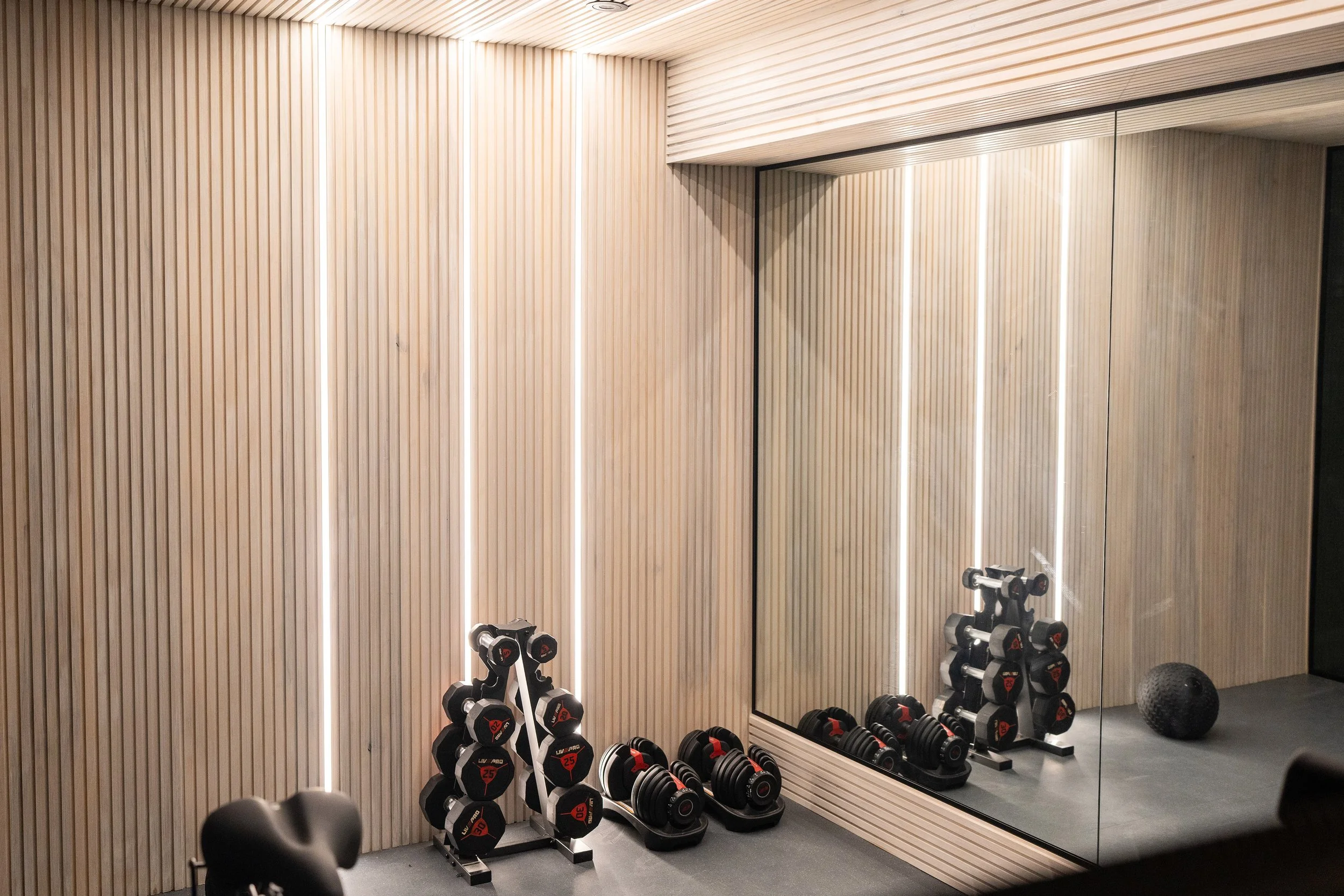 A gym corner with dumbbells on rack, workout bands, a textured wall, a large mirror, and a black exercise ball.