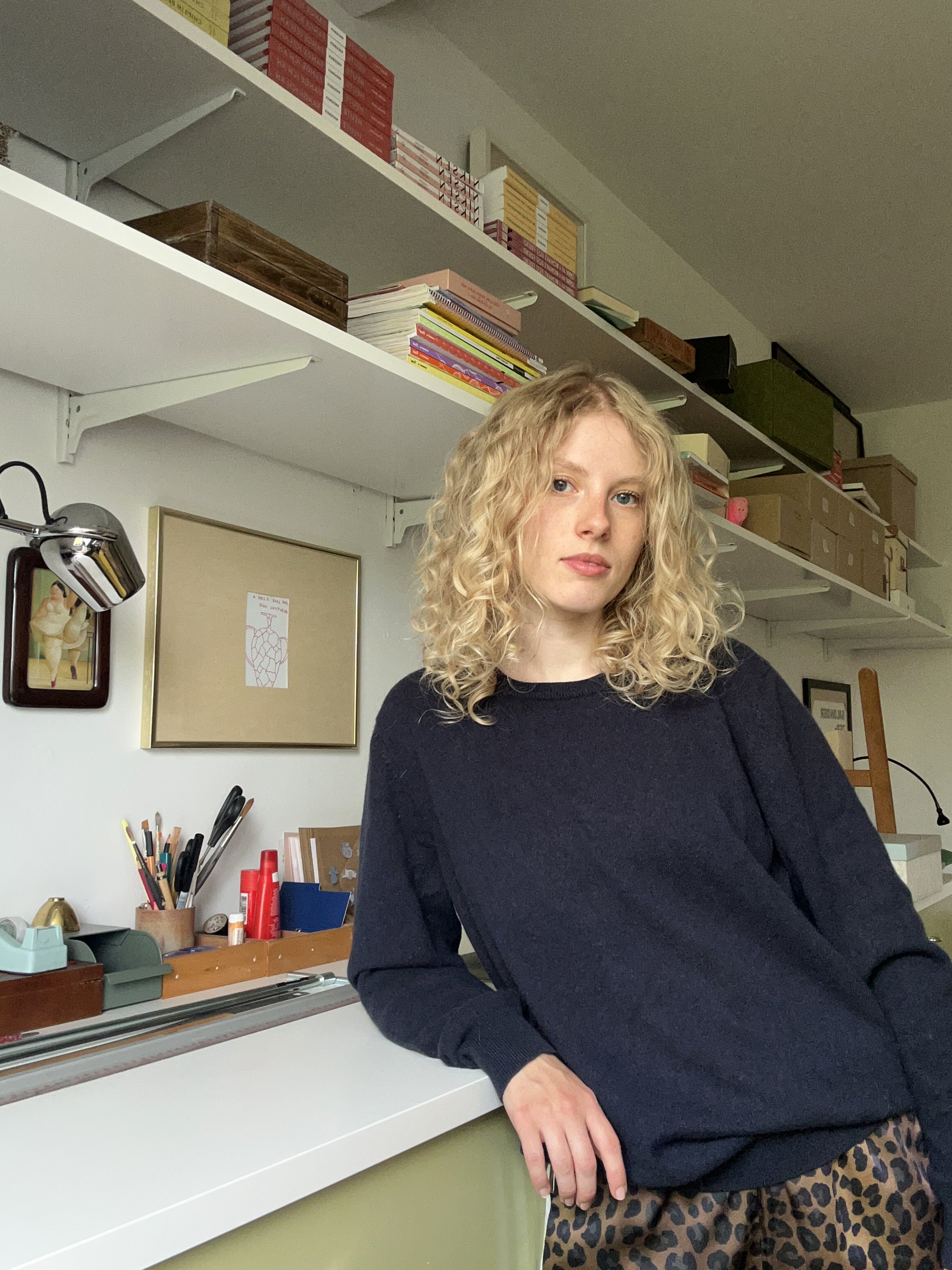 A young woman with curly blonde hair and fair skin standing in a room with bookshelves and office supplies, wearing a black sweater and leopard print pants.