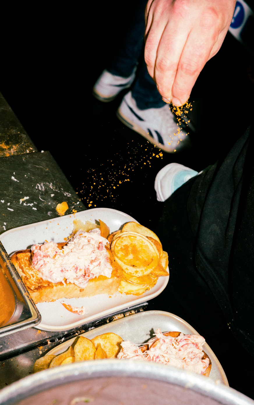 Close-up of a chef’s hand sprinkling seasoning over lobster rolls with crisps, captured with bold detail by London food photographer Edward Howell.