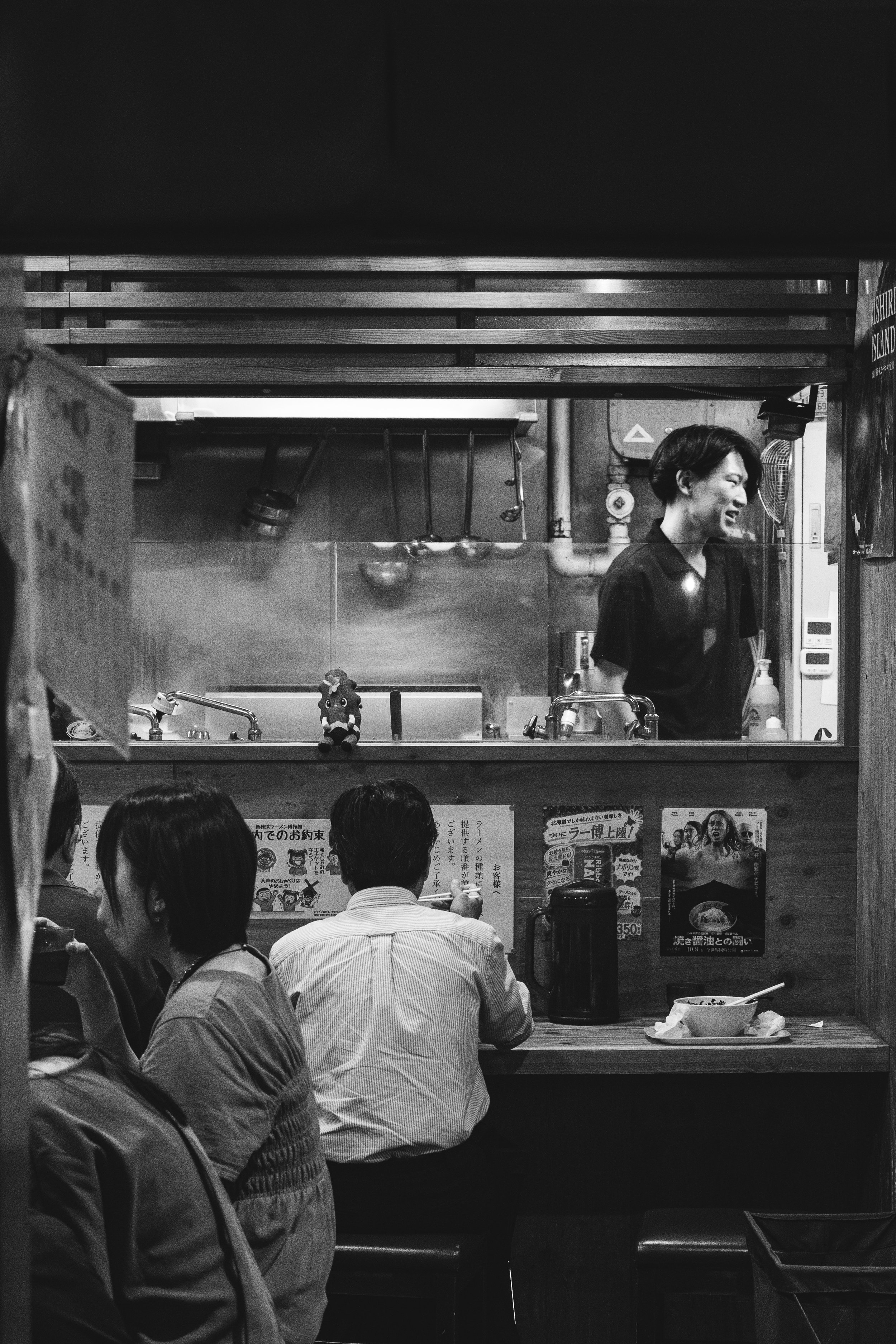 A black and white photo of a restaurant or food stall, with a woman behind the counter smiling and laughing. Several customers are sitting at the counter eating. The scene appears lively and casual.
