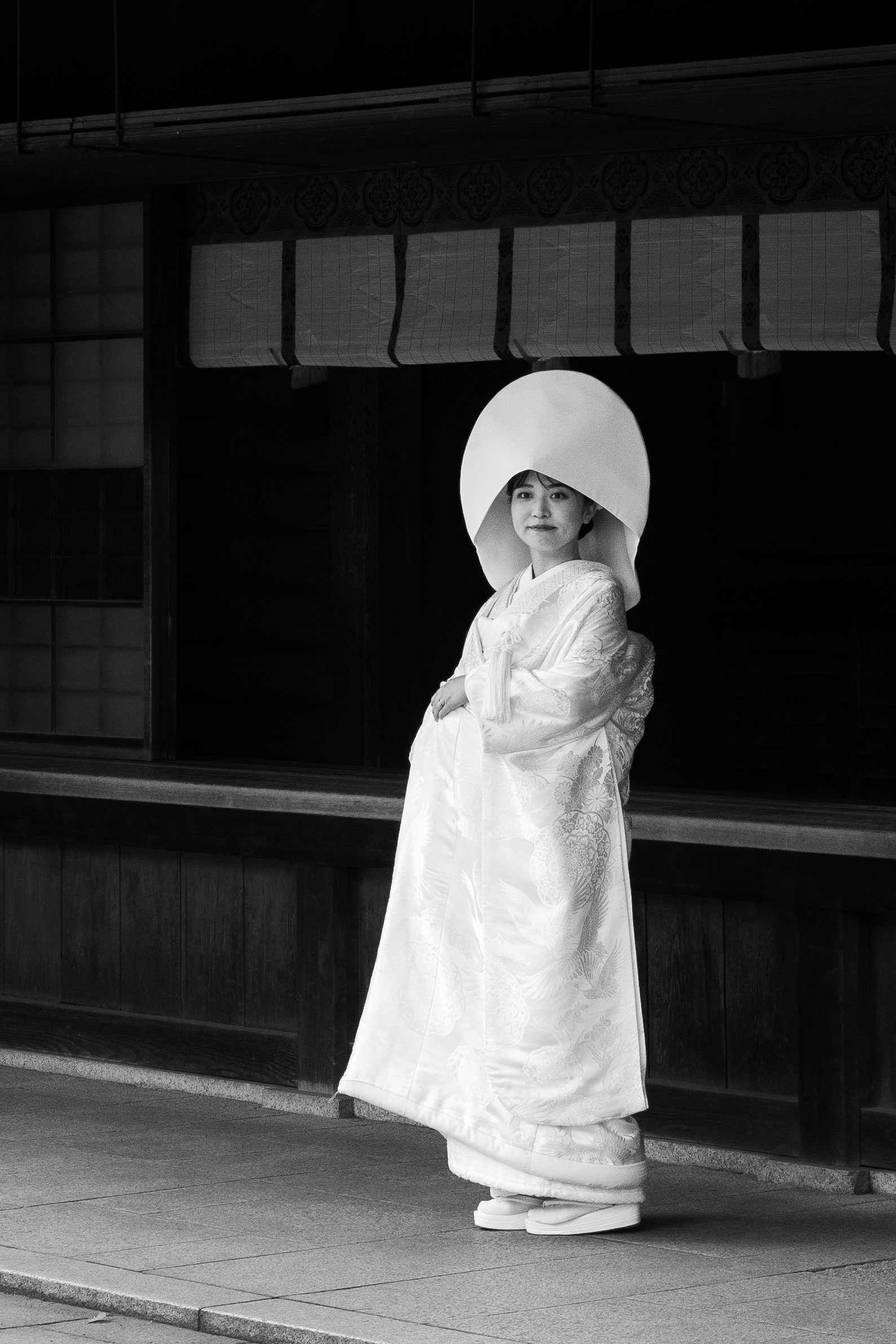 A woman dressed in traditional Japanese bridal kimono and wearing a large white hood, standing on a sidewalk in front of a building with sliding doors.