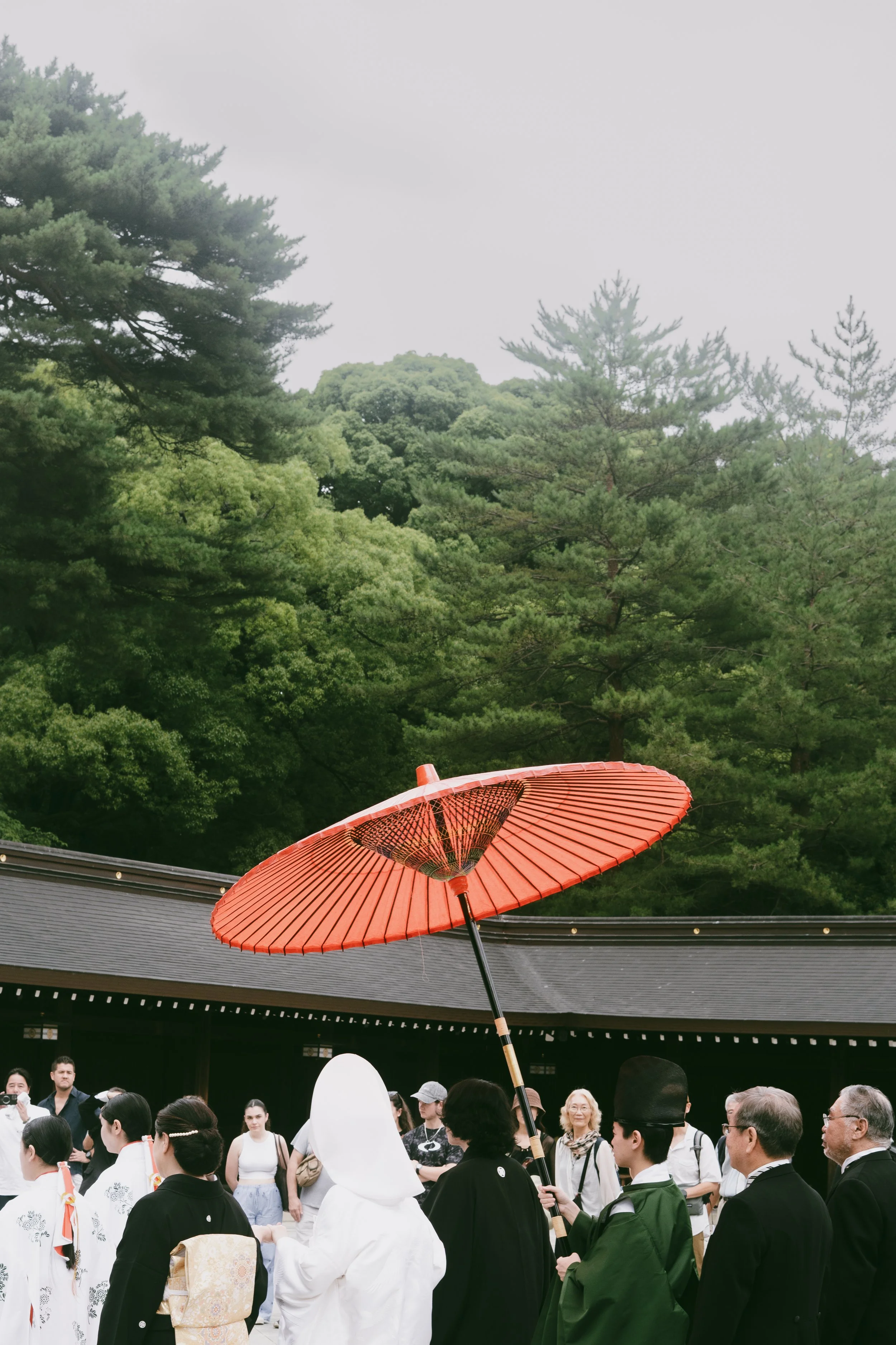 Crowd of people gathered outdoors with a traditional red umbrella held above them, green trees in the background.