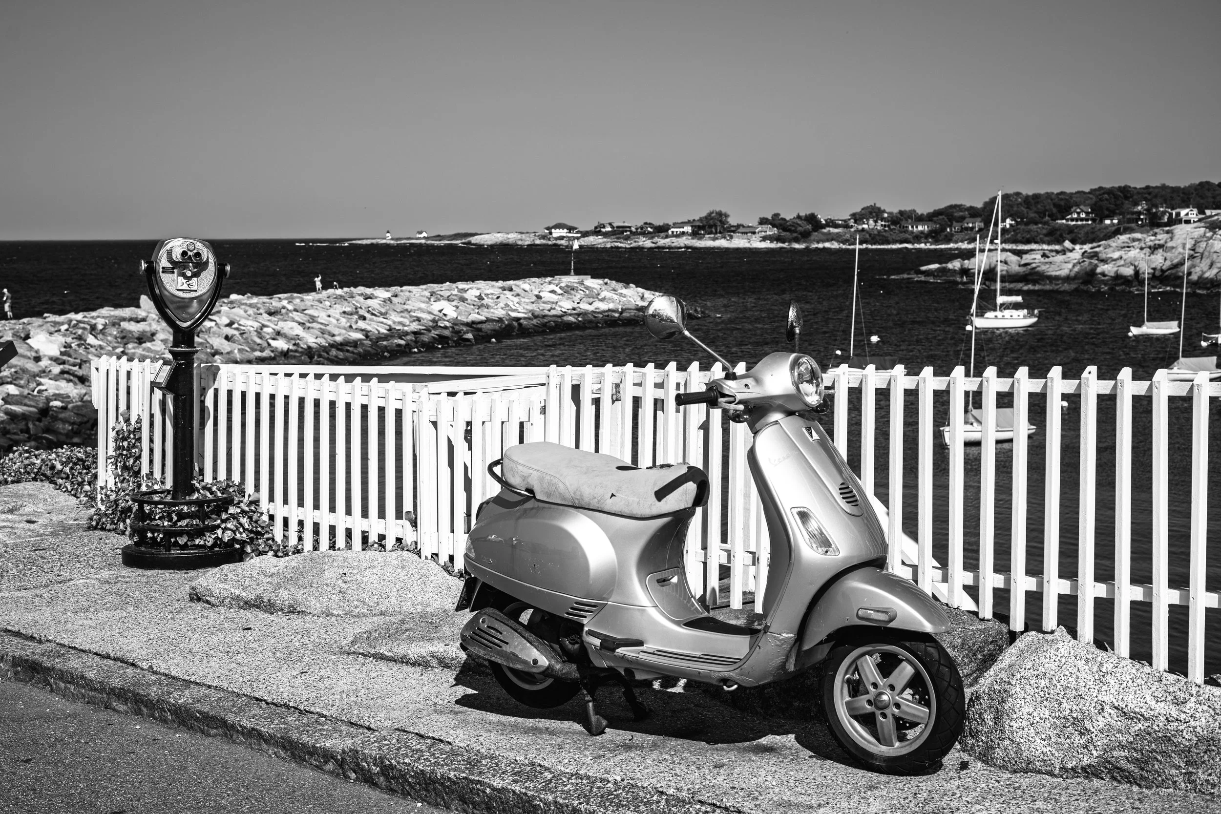 Black and white photo of a scooter parked on a sidewalk near a white fence with a view of a rocky shoreline, sailboats, and houses in the background.
