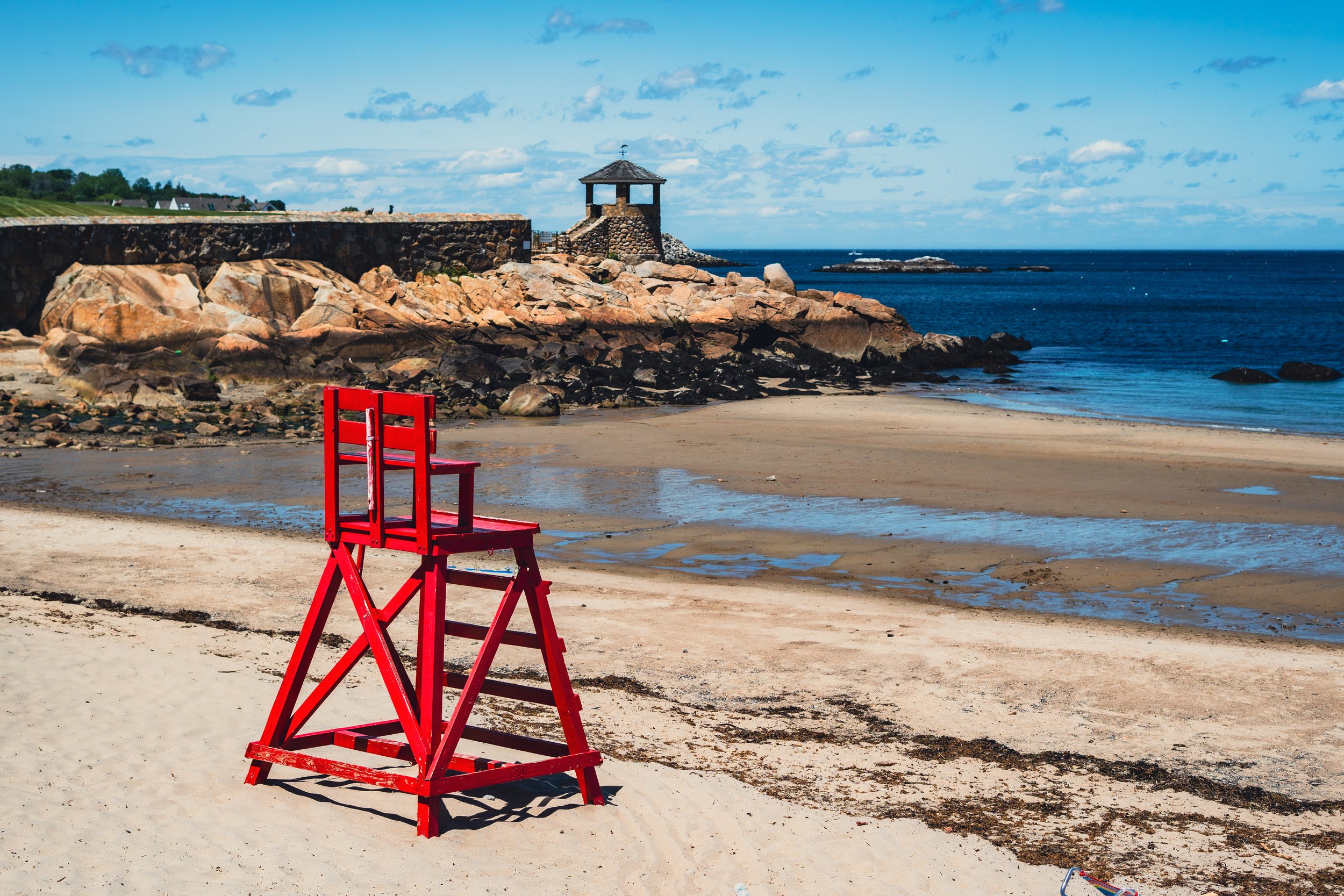 Red director's chair on sandy beach with rocks, ocean, and partly cloudy sky in the background.