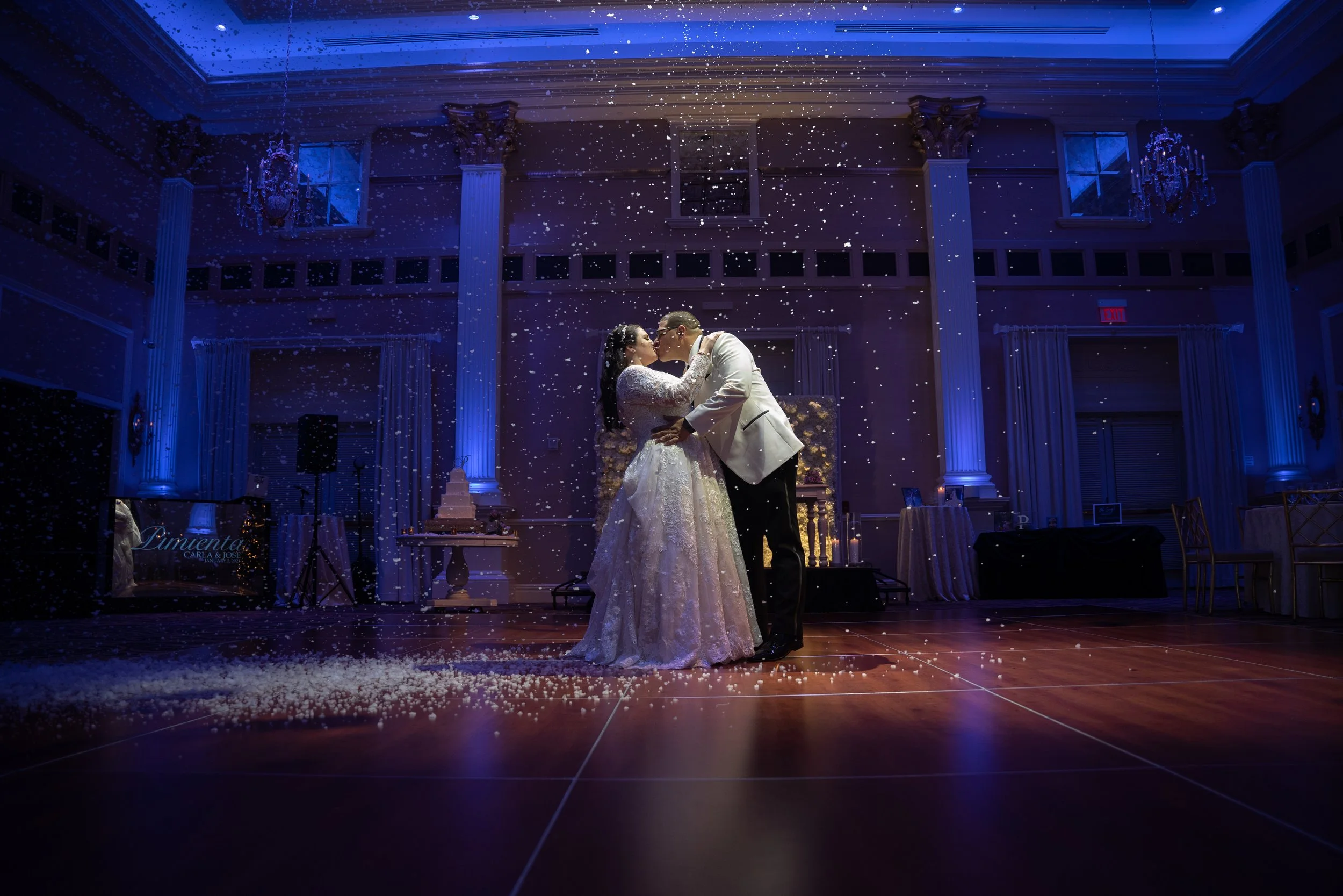 A bride and groom share a kiss on the dance floor at their wedding reception, with falling confetti and candlelight in the background.