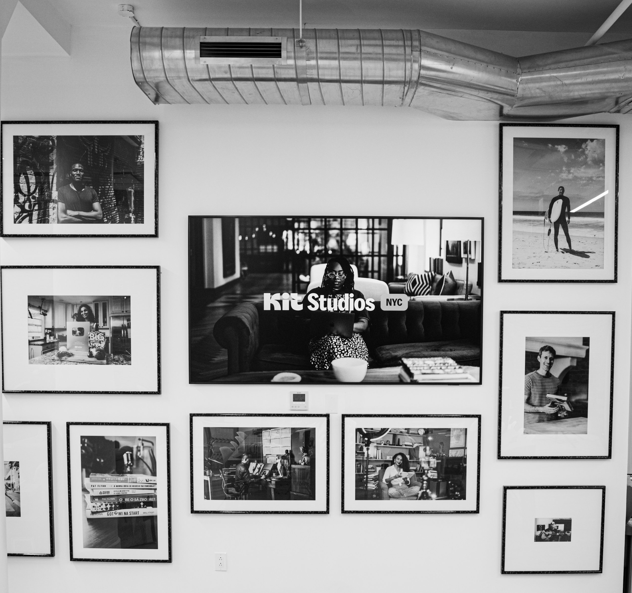 Black and white photo wall with framed pictures in an art studio titled Kit Studios NYC, featuring portraits of people and a surfing scene, with a woman sitting on a couch in the center.