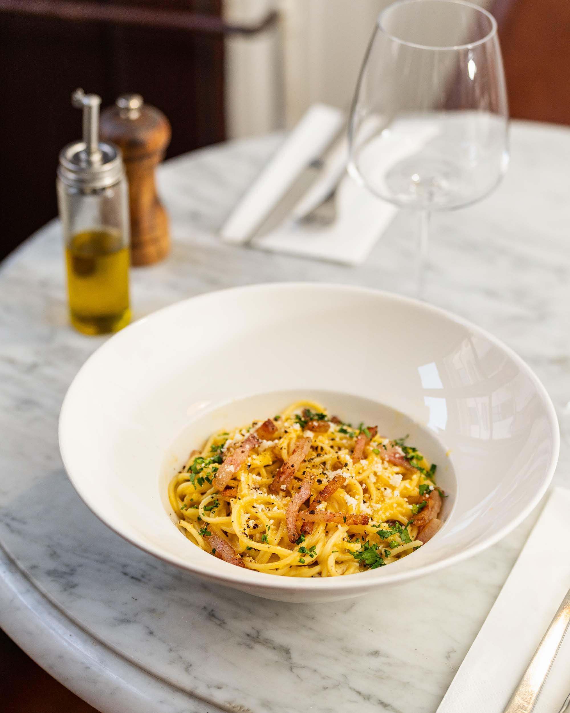A bowl of spaghetti pasta with bacon pieces, garnished with herbs on a marble tabletop, with a glass of water, a salt shaker, and a pepper mill in the background.