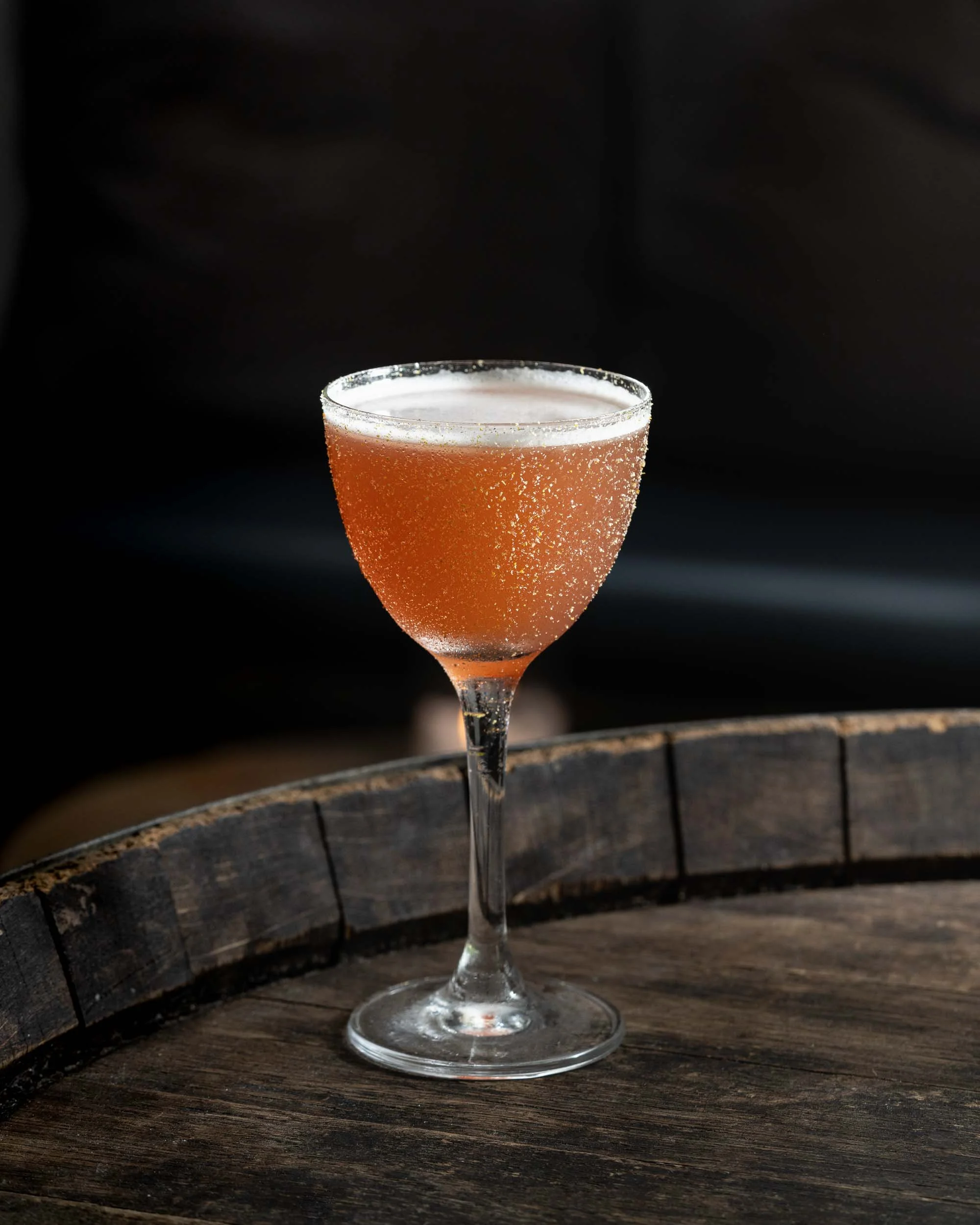 A martini glass filled with a pinkish-orange beverage, garnished with sugar or salt on the rim, set on a wooden surface with a dark background.