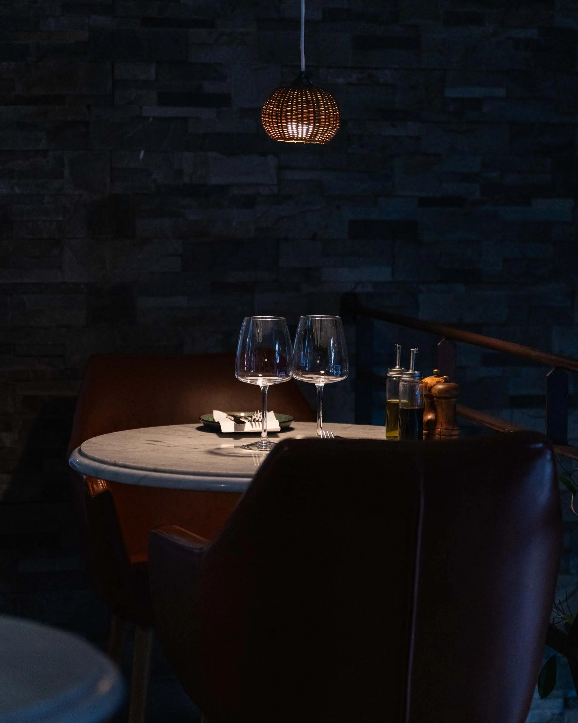 Dimly lit dining setting with a white marble table, two empty wine glasses, and a dark background. A warm-toned woven pendant light hangs from the ceiling, illuminating part of the table. A plate with cutlery, a small bottle of olive oil, and a pepper grinder are visible.