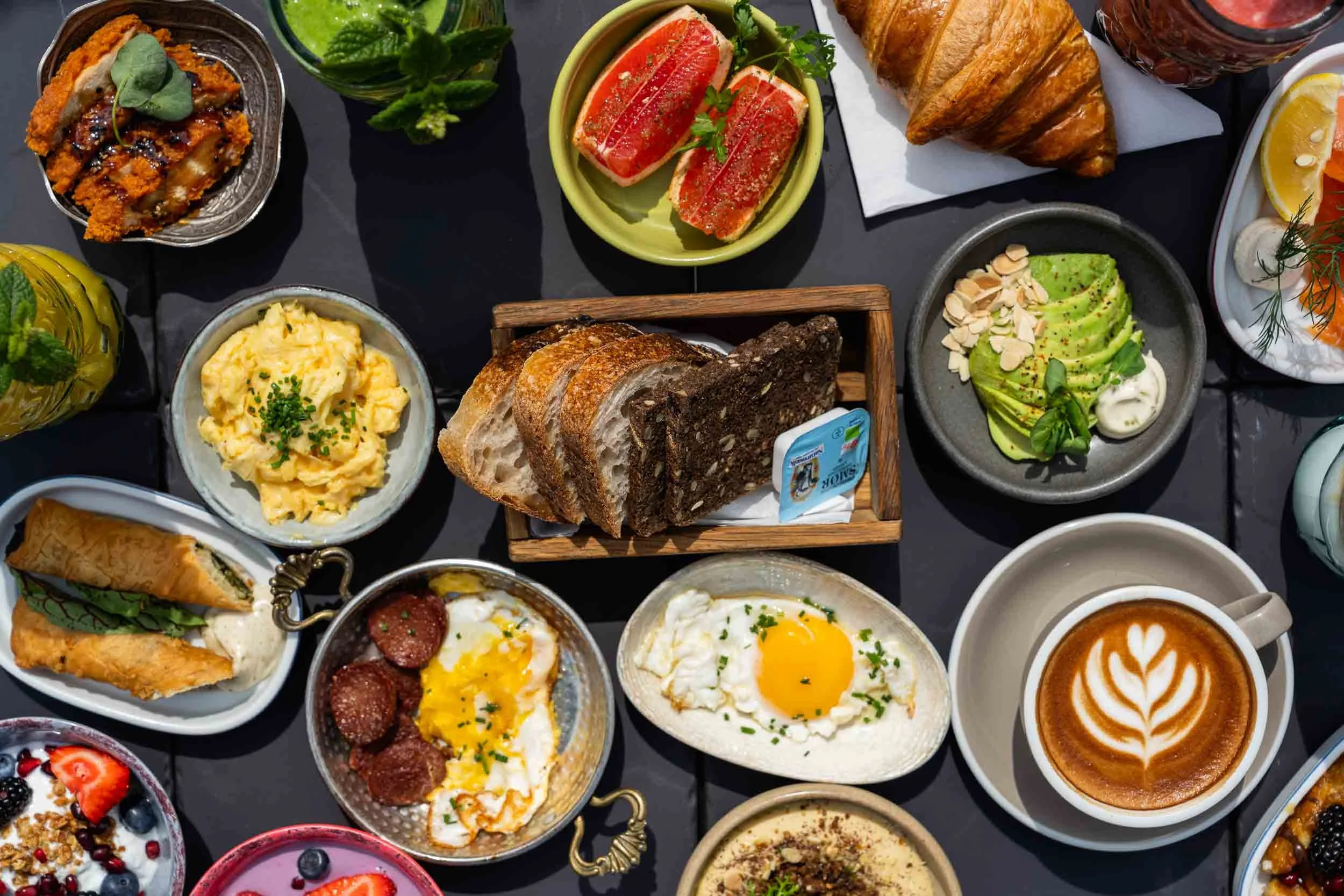 A variety of breakfast foods including scrambled eggs, fried eggs, croissants, slices of bread, avocado slices, strawberries, blueberries, and beverages on a dark table.