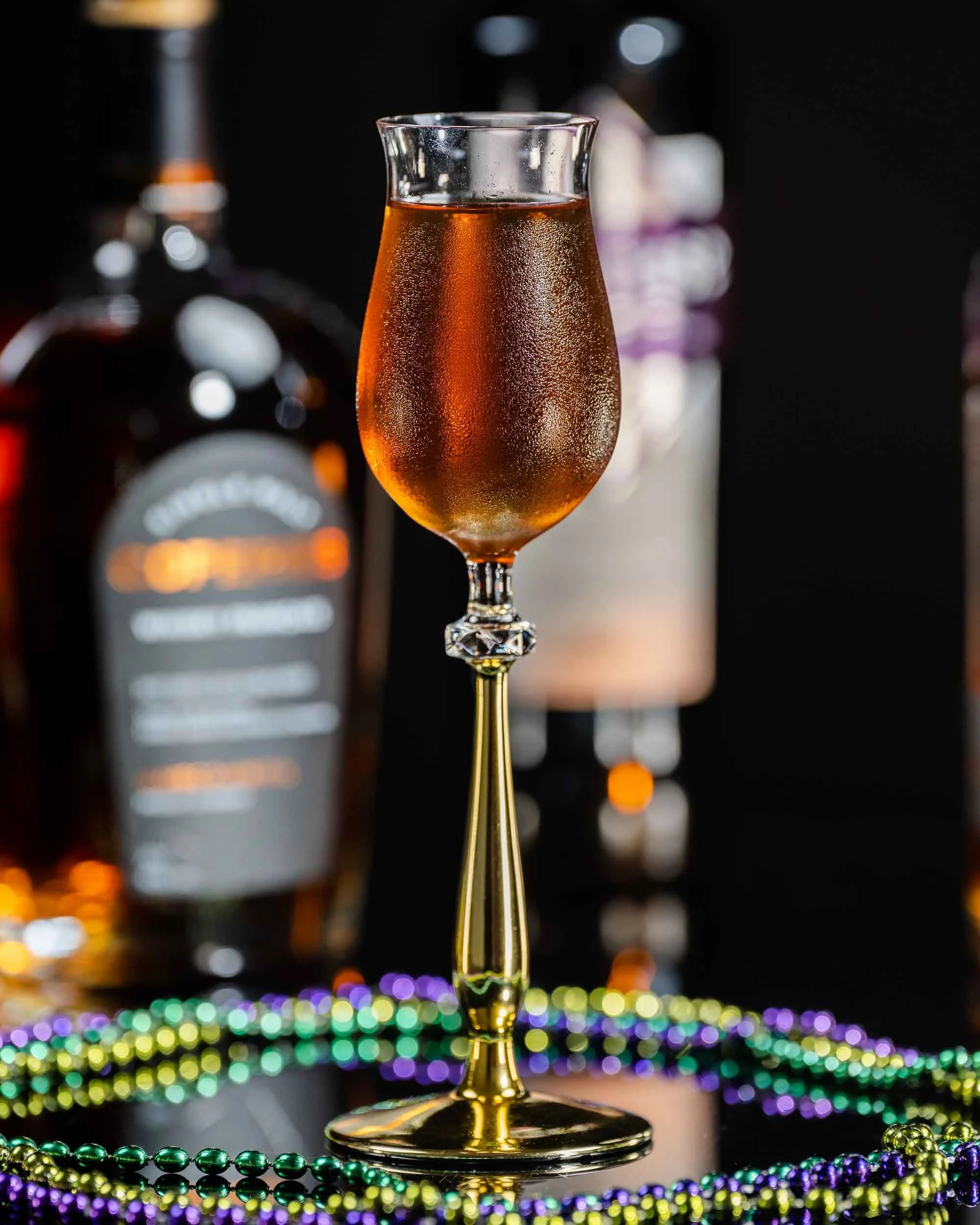 A champagne flute filled with sparkling golden-colored beverage, decorated for celebration with colorful beads on a black surface, against a dark background with blurred bottles.