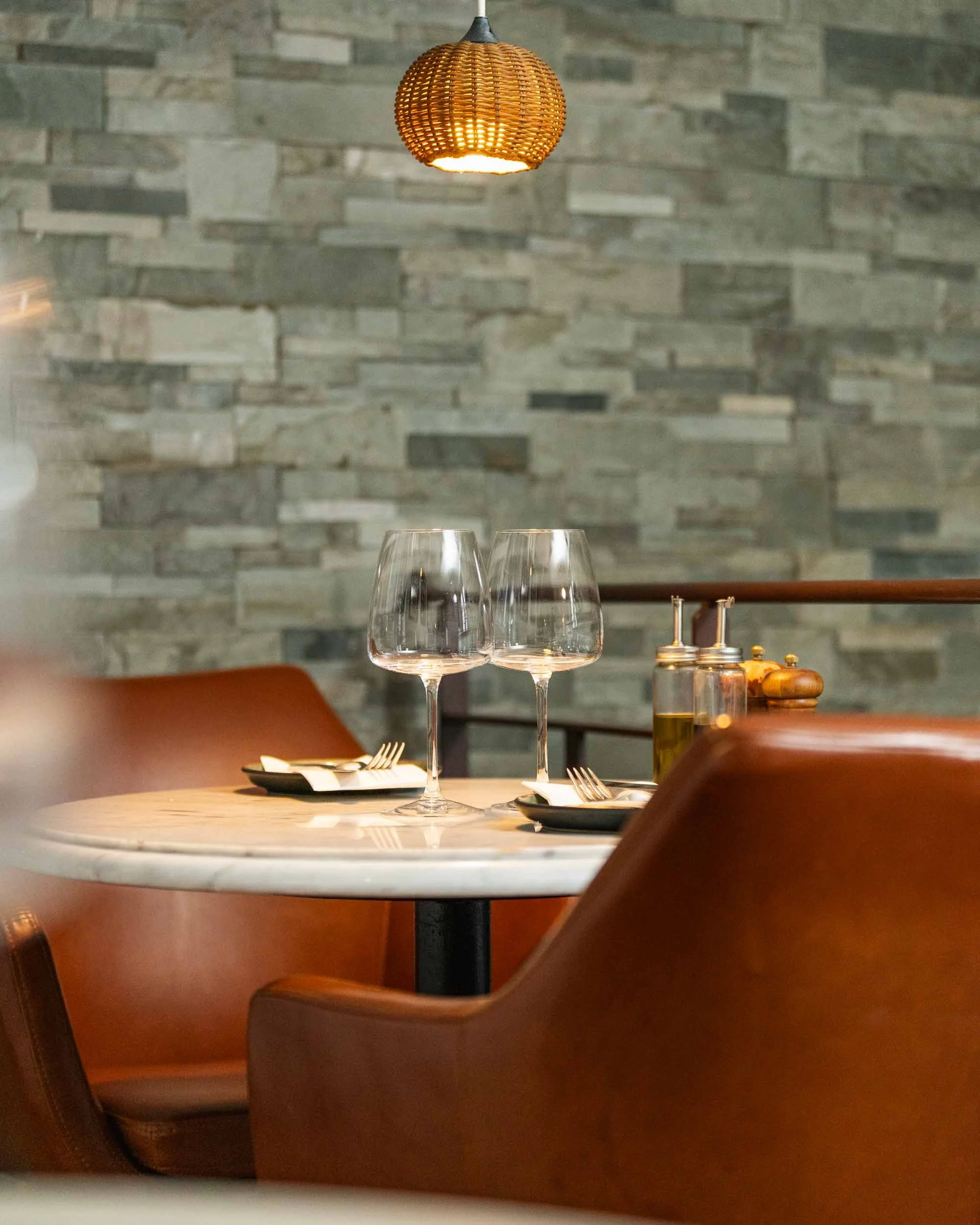 A table set with two empty wine glasses, a black plate, and silver utensils, in a restaurant with a stone brick wall and a hanging wicker light in the background.