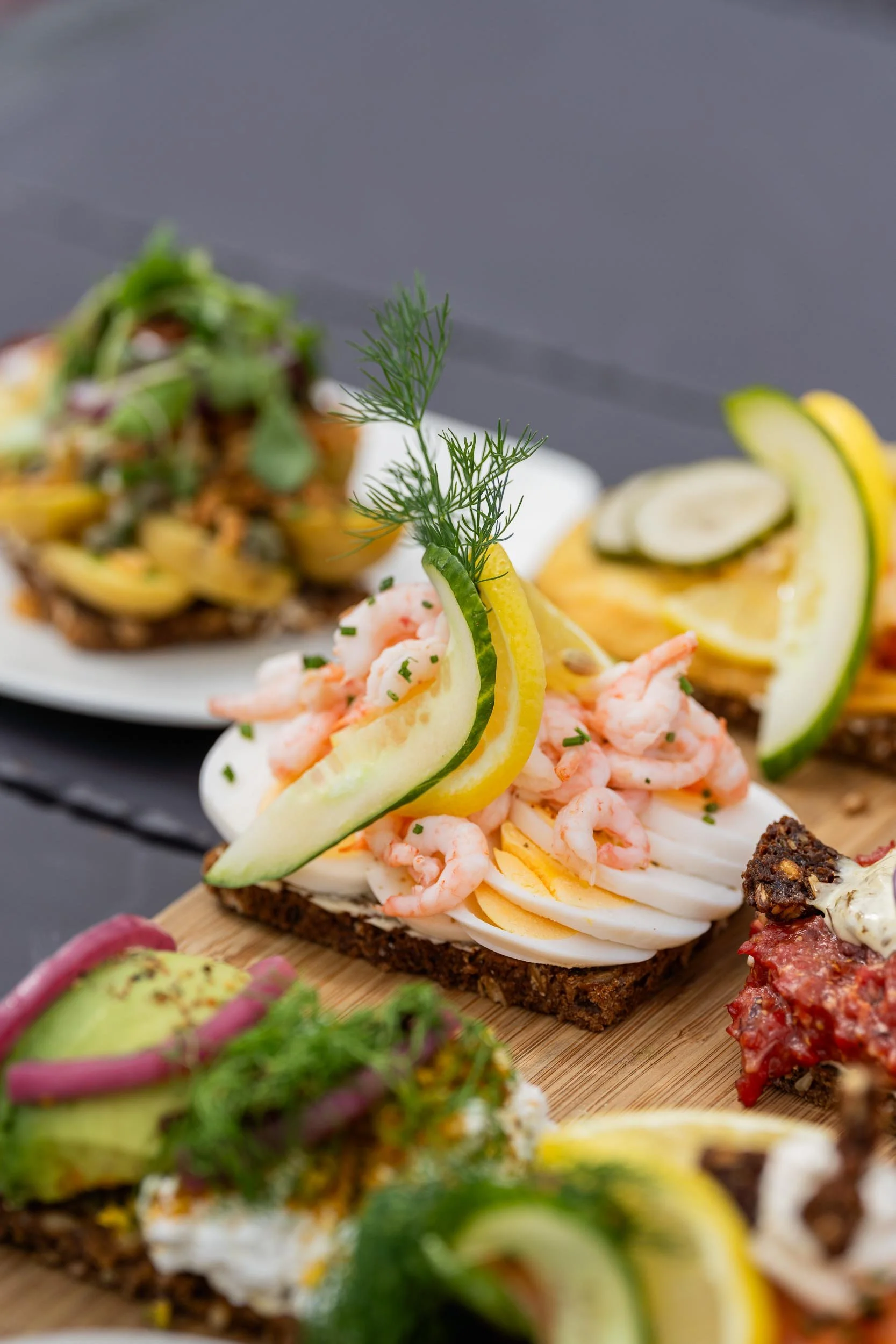 Close-up of assorted open-faced sandwiches with toppings including shrimp, lemon slices, cucumber, dill, and various garnishes on a dark surface.