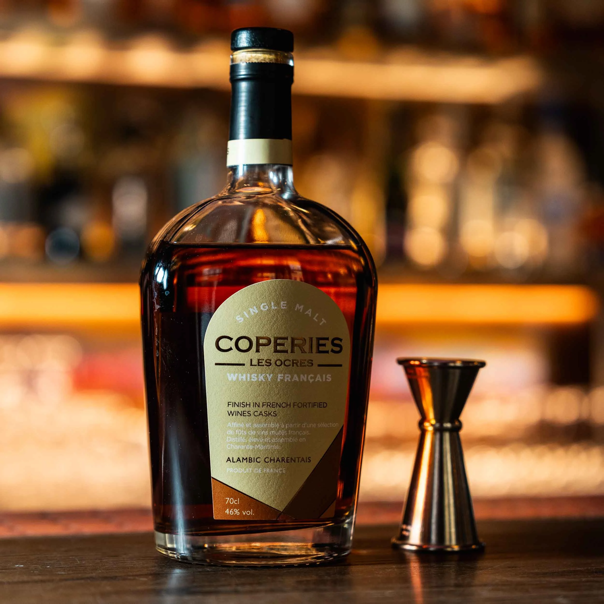 Close-up of a bottle of Coperries Les Ogres whiskey with a blurred background of bar shelves.