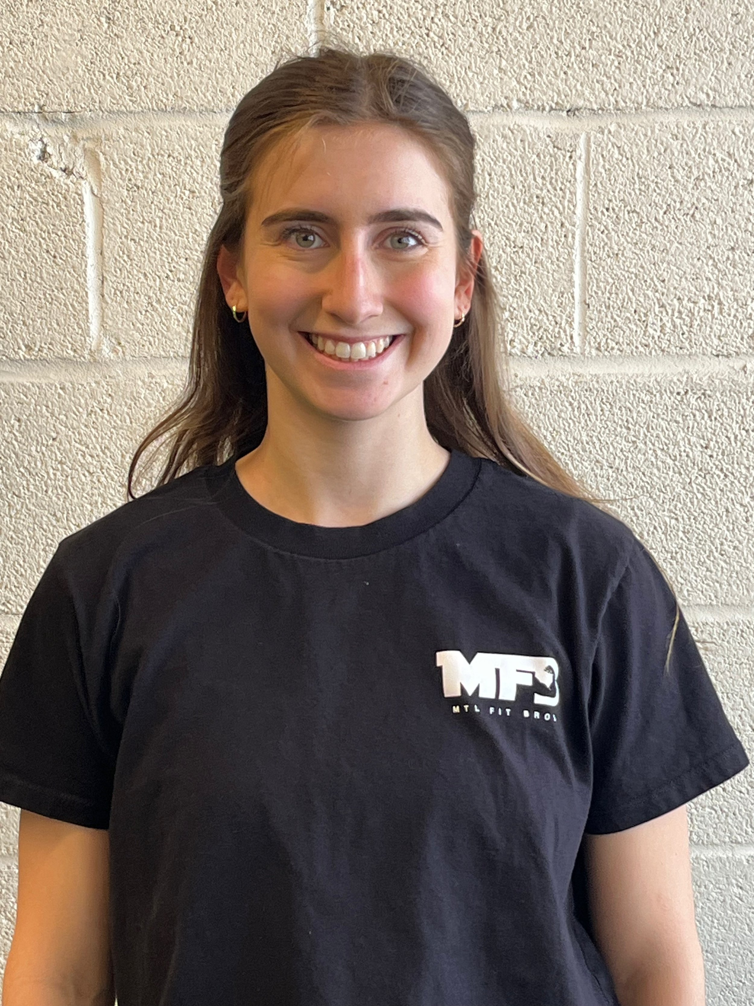 A young woman with shoulder-length light brown hair smiling, wearing a black sweatshirt with white text and logo, standing in front of a plain black wall.