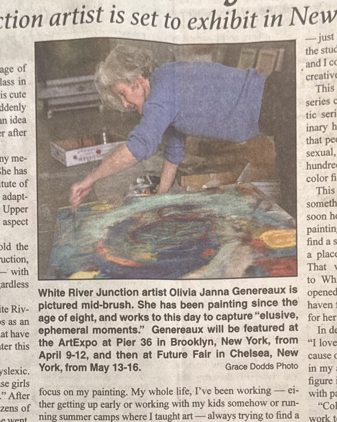 A woman, Olivia Janna Genereaux, with short gray hair wearing a blue shirt and brown pants leaning over a large colorful abstract painting on a table, working with a paintbrush.