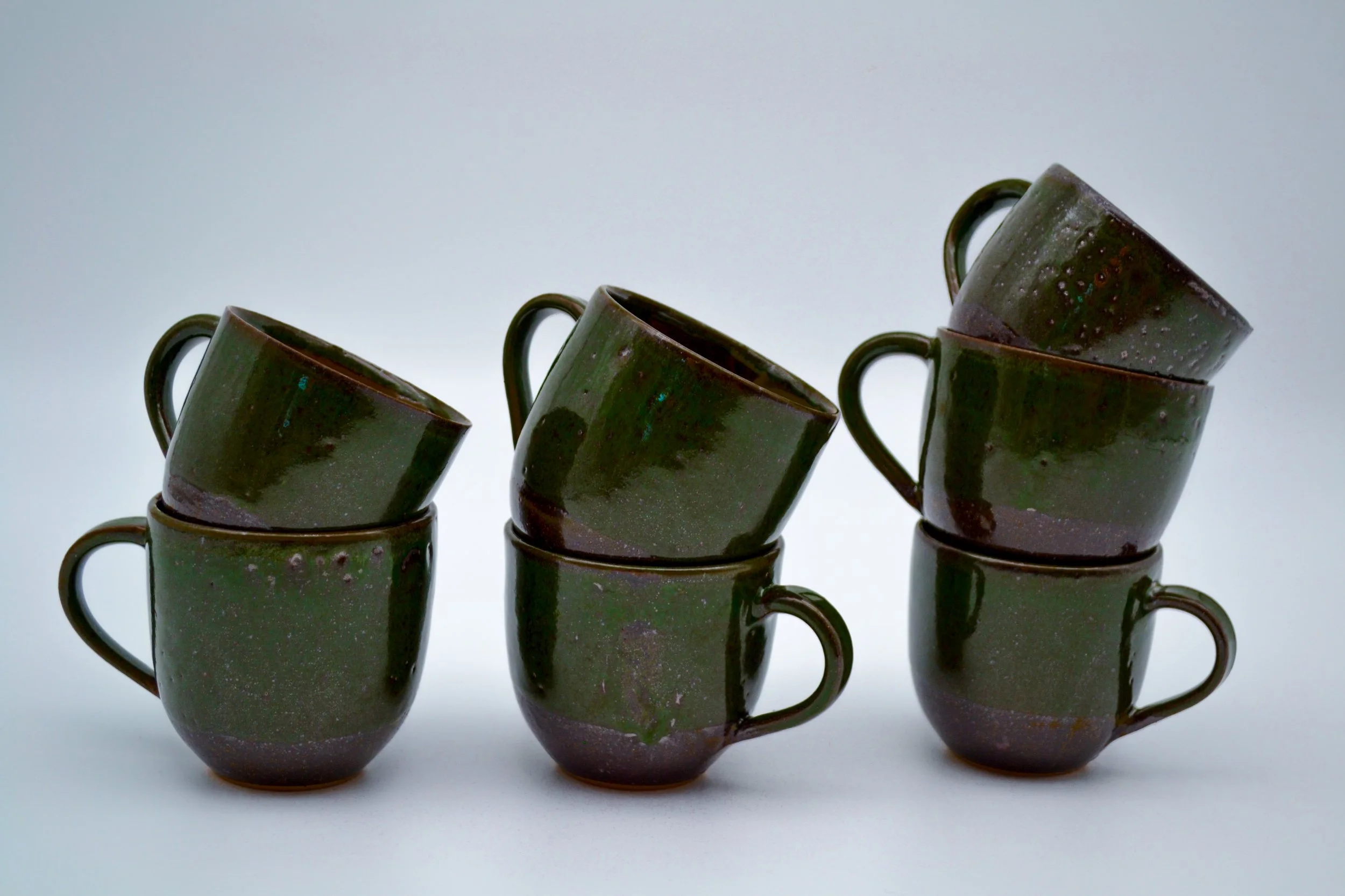 Six dark green ceramic mugs with a speckled glaze, stacked in two columns of three against a light gray background made by artist Allison Genereaux.