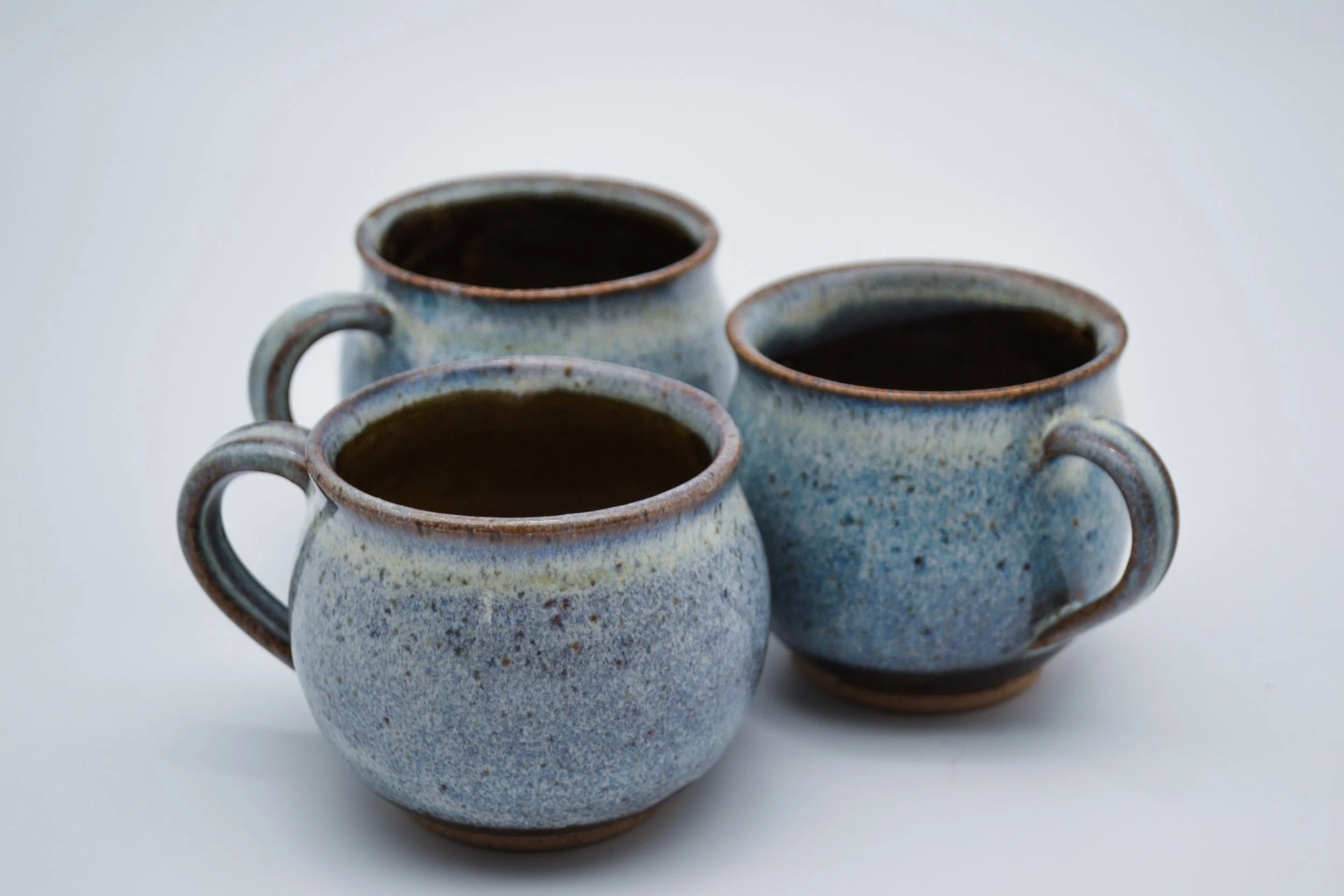 Three ceramic mugs filled with dark liquid arranged in a cluster on a white surface against a plain background made by artist Allison Genereaux of Alcove Ceramics.