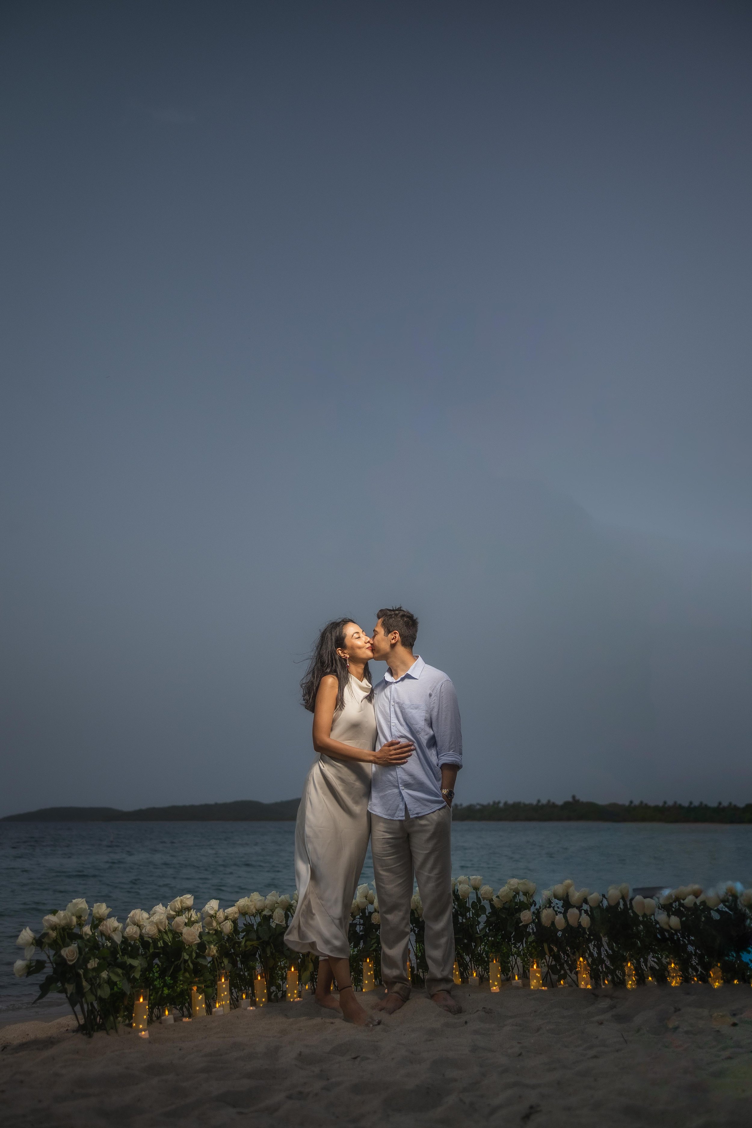Brijend and Sumi share a kiss beside a candlelit white rose arrangement overlooking the Caribbean Sea during their destination marriage proposal in Puerto Rico.