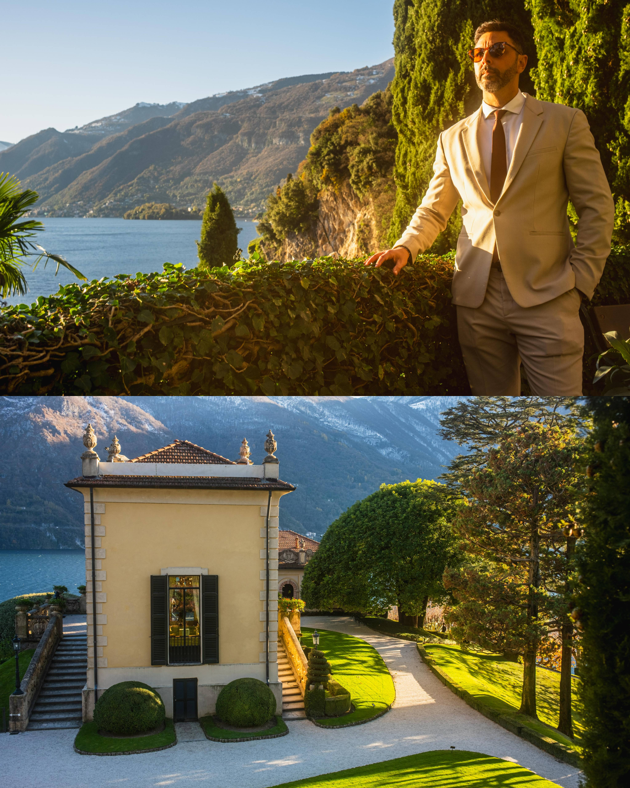 Man in beige suit standing beside cypress trees at Villa del Balbianello overlooking Lake Como mountains in Italy.