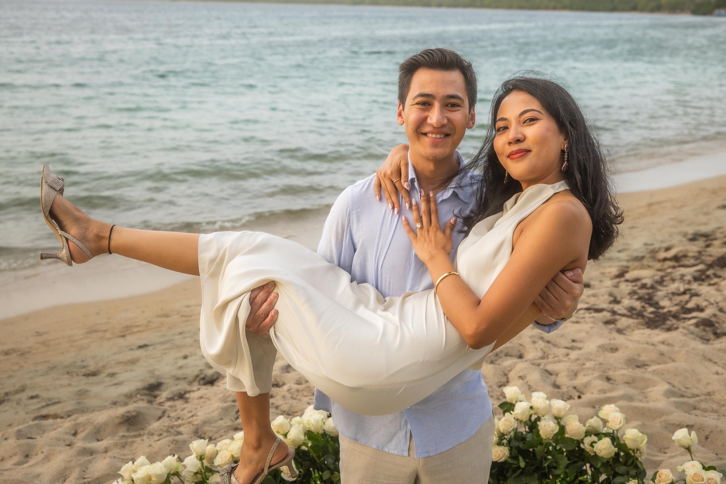 Brijend carries Sumi in his arms on the beach surrounded by white roses after their destination marriage proposal in Puerto Rico.