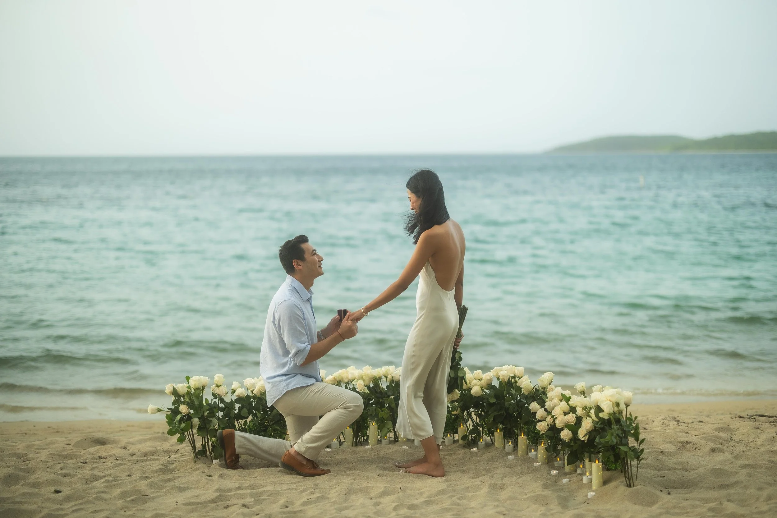 Romantic beach proposal photograph of Brijend kneeling before Sumi as he presents her with an engagement ring surrounded by white roses in Puerto Rico