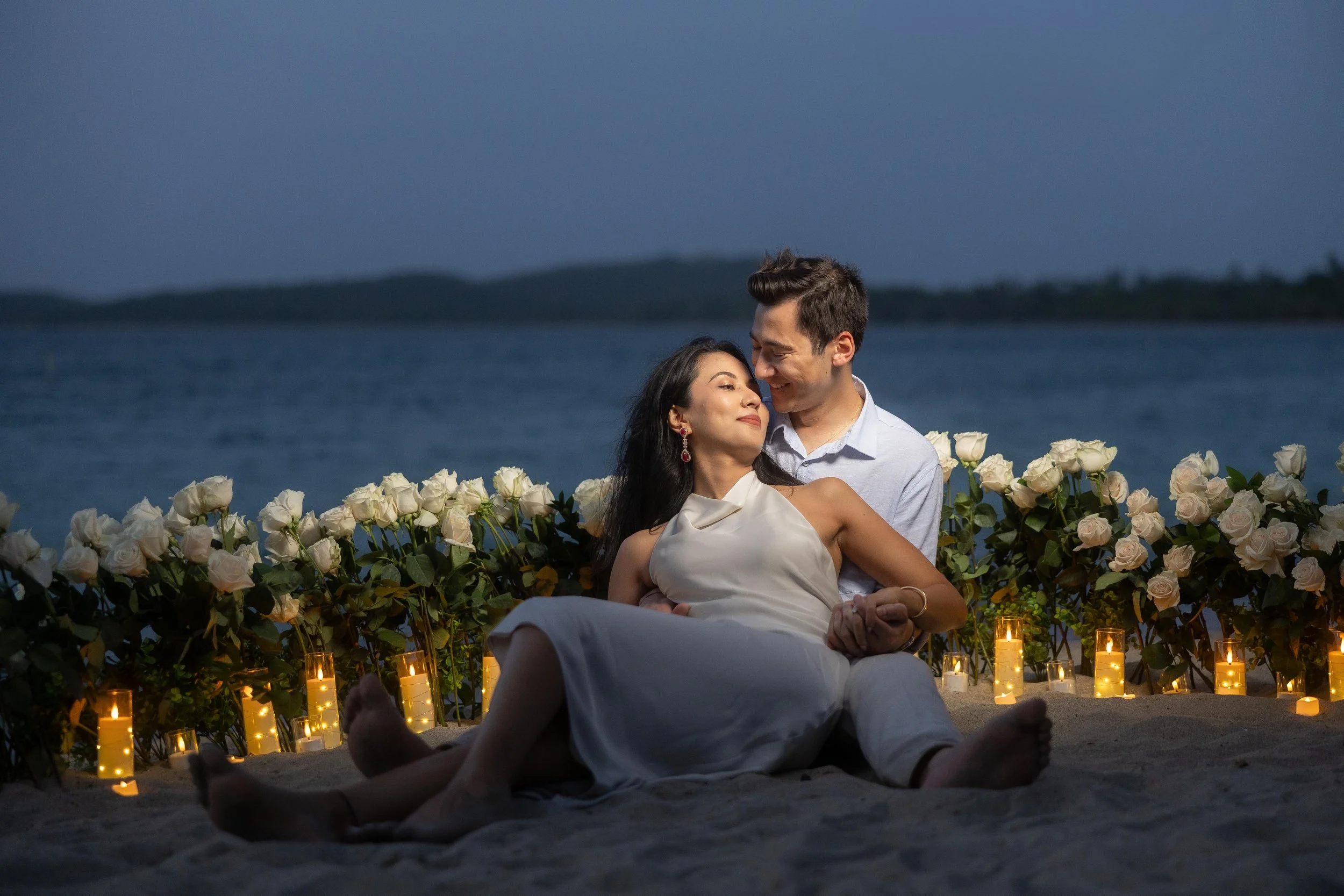 Brijend and Sumi sit together on the beach surrounded by white roses and candlelight after their romantic marriage proposal in Puerto Rico.