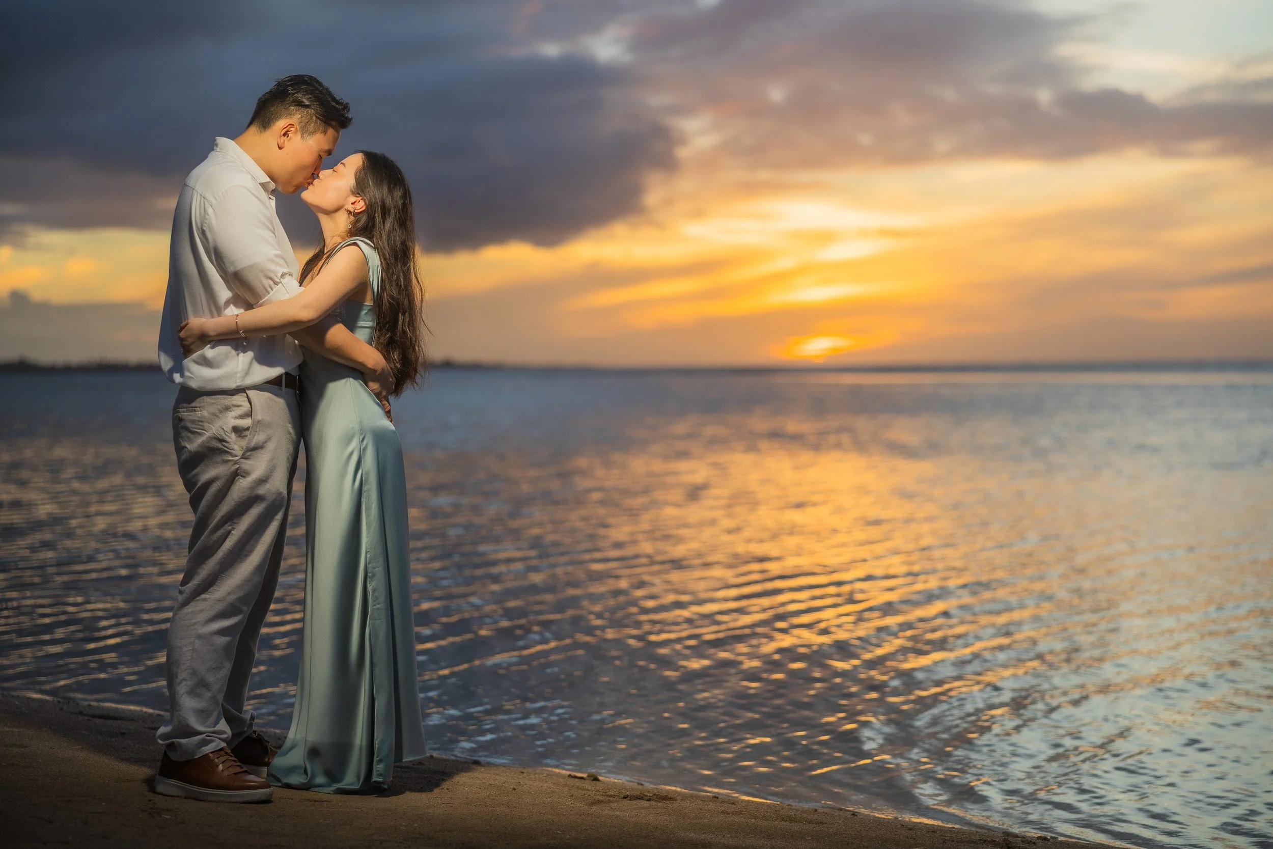 Newly engaged couple embracing and sharing a kiss along the shoreline at Hyatt Regency Grand Reserve in Puerto Rico. Golden sunset colors reflect across the water, creating a romantic backdrop for their luxury marriage proposal session.