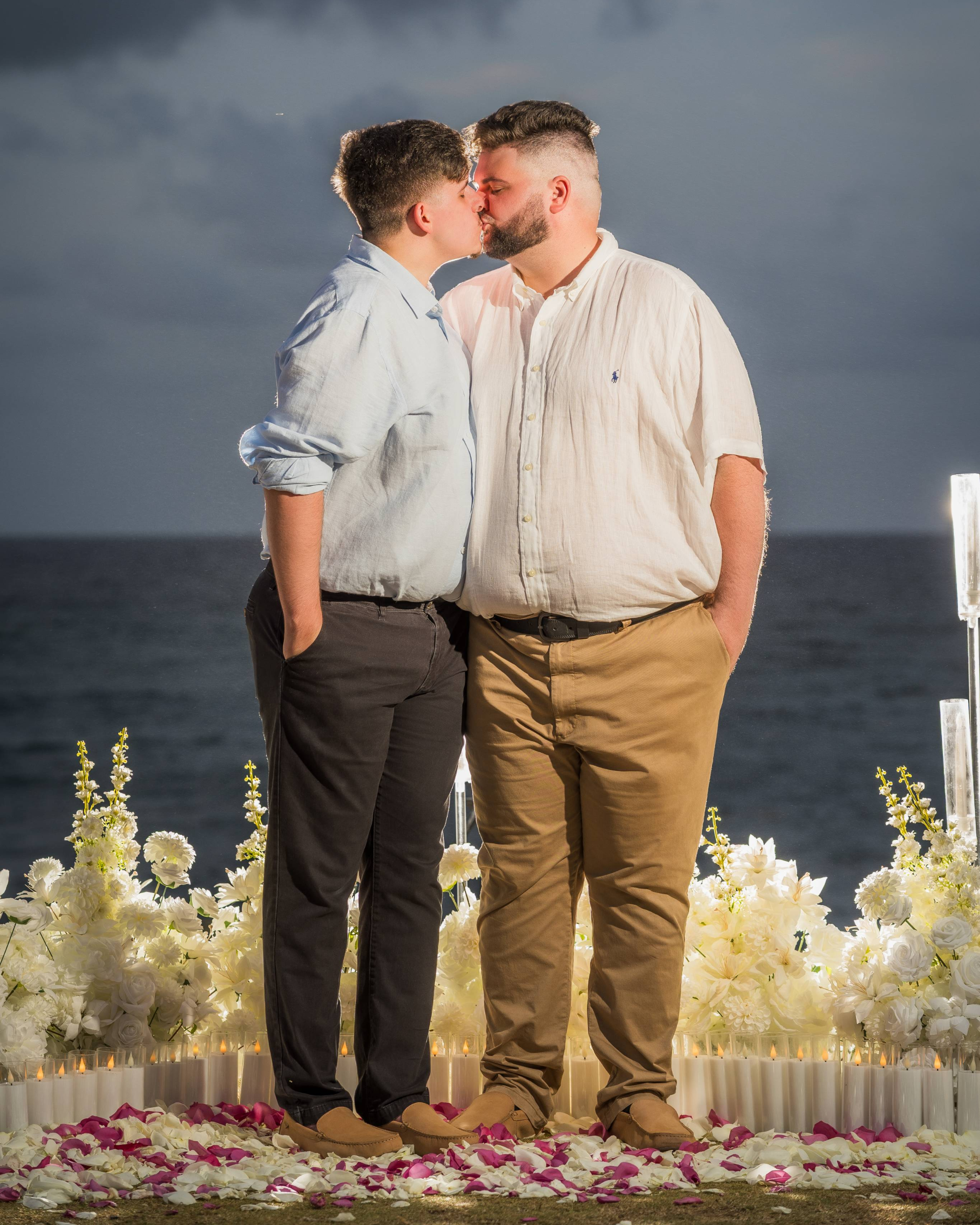 Two men share their first kiss as an engaged couple beside an elegant oceanfront proposal setup decorated with white flowers and candles at Escambrón Beach in Puerto Rico.
