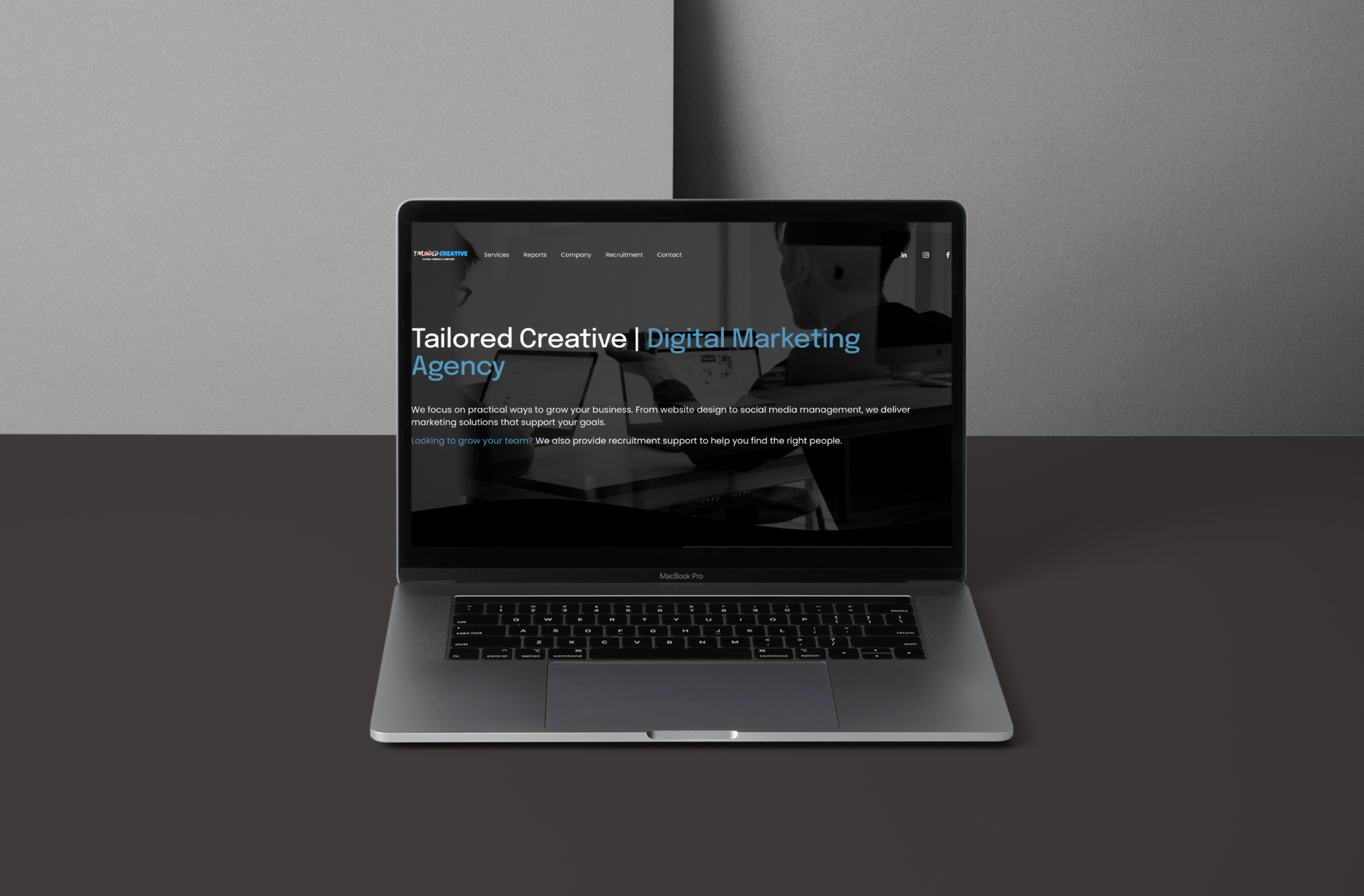 A laptop on a round wooden table displaying a website for Tailored Creative, a digital media agency, with a background of black and wooden shelving