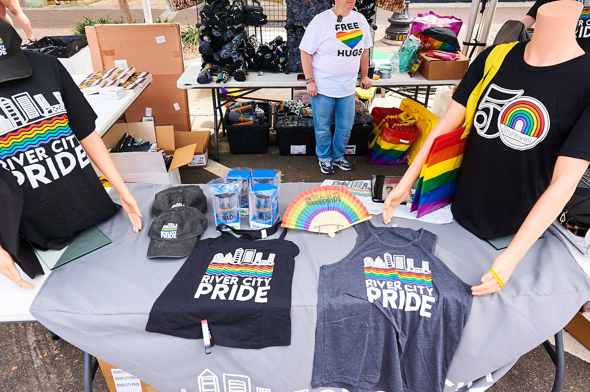 Display of River City Pride merchandise, including T-shirts, hats, mugs, and rainbow-themed items at an outdoor vendor booth. A person wearing a 'Free Hugs' shirt is in the background.