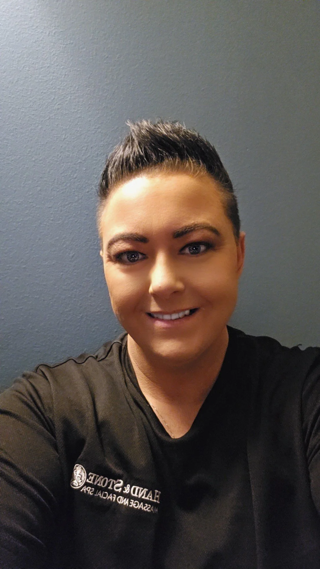 A woman taking a selfie against a plain blue wall. She has short, dark hair styled upward, light makeup, and is wearing a black shirt with white text and logo that says 'HAND & STONE MASSAGE AND FACIAL SPA'.