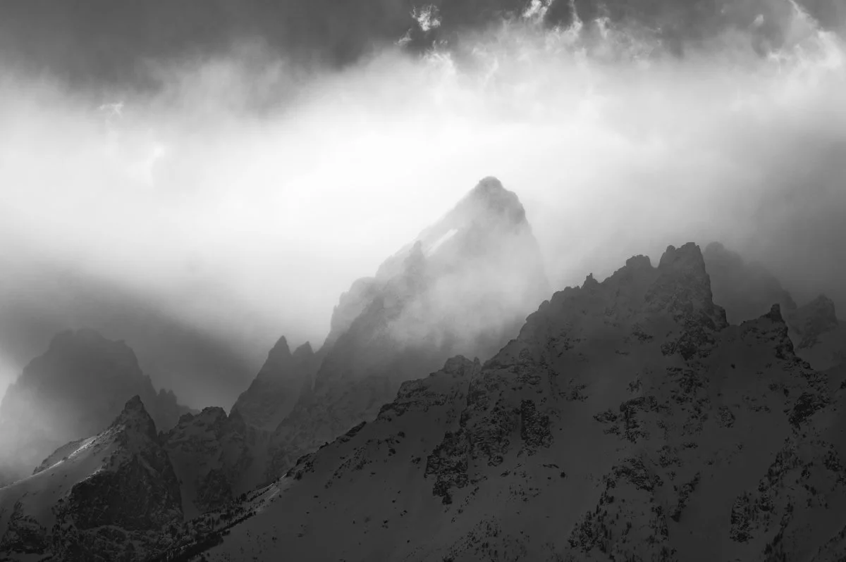 Black and white photograph of rugged mountain peaks shrouded in clouds and mist.