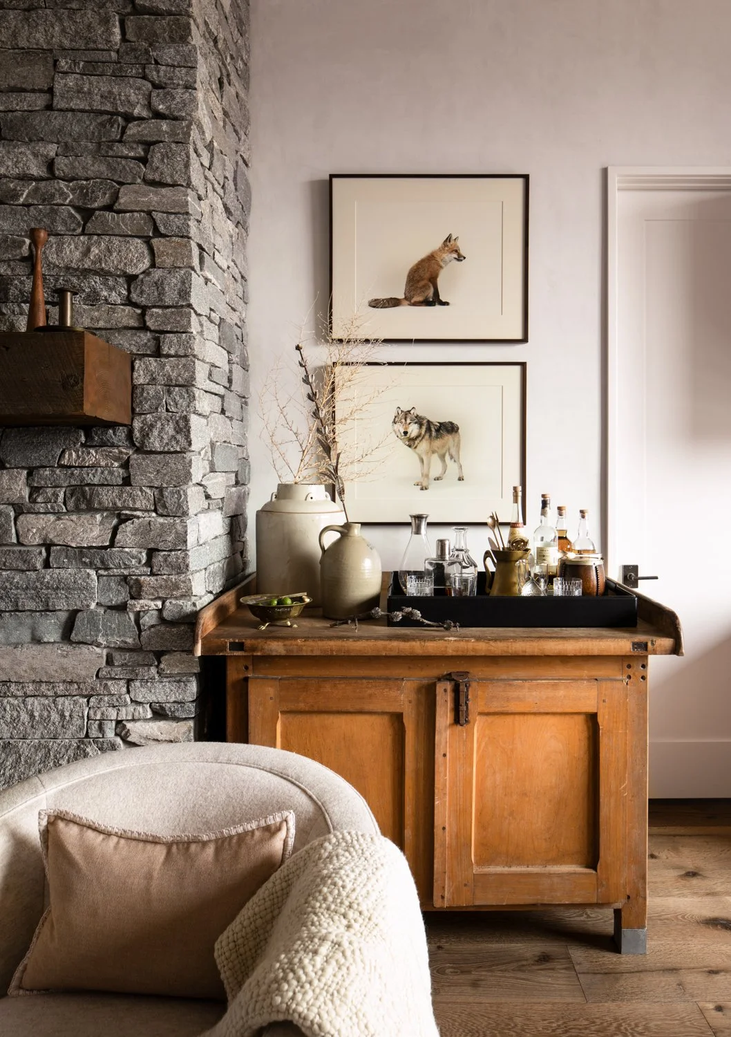 Interior room with stone wall, wooden sideboard, decorative vases, framed animal artwork, and a cozy armchair with a pillow and knitted throw.