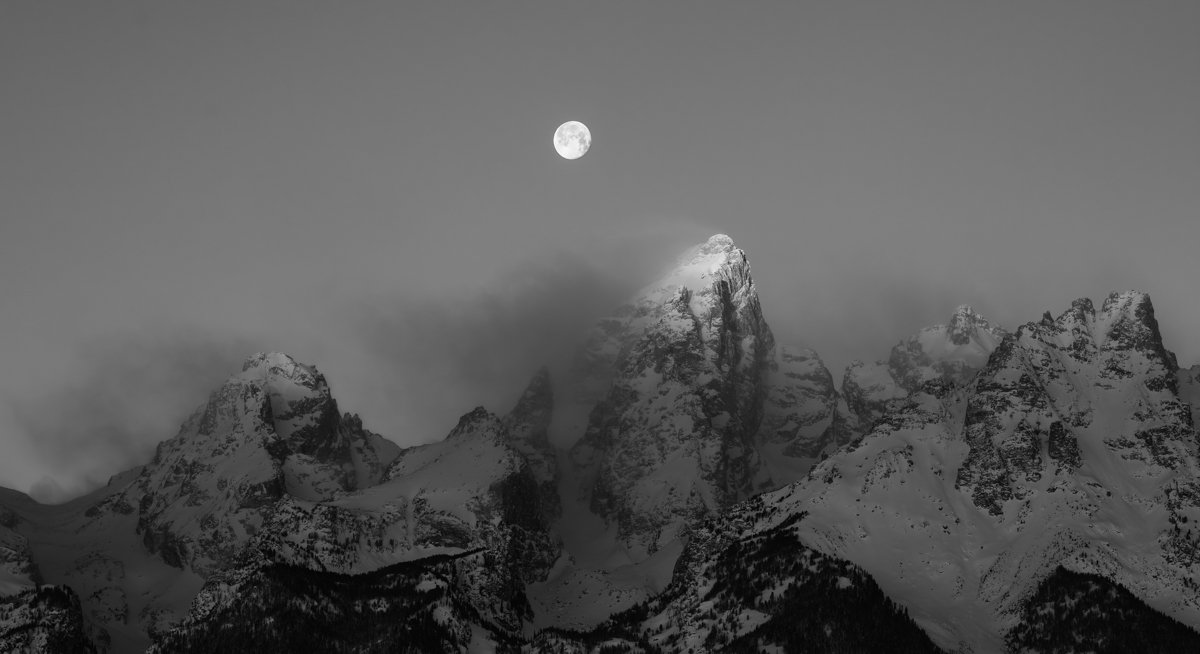 Nighttime landscape of snow-covered mountains with a visible full moon in the sky.