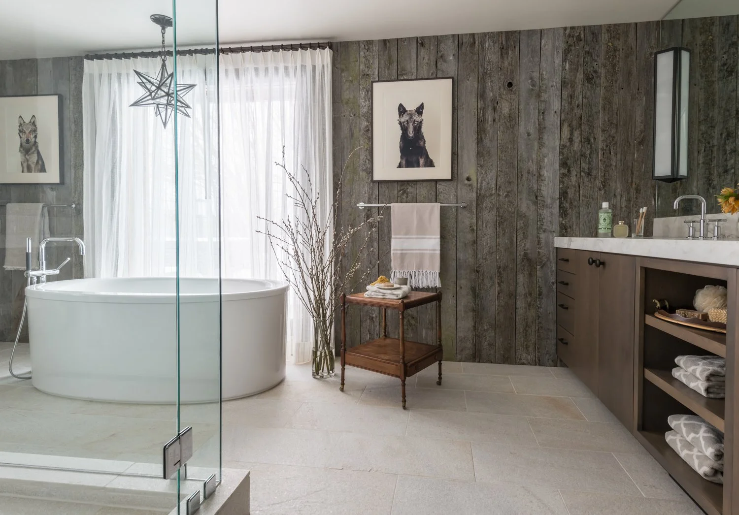 Modern bathroom with a freestanding bathtub, glass shower enclosure, wooden vanity with a sink, and framed animal artwork on a rustic wood-paneled wall. Decor includes white towels, dried branches in a glass vase, and small bathroom essentials.