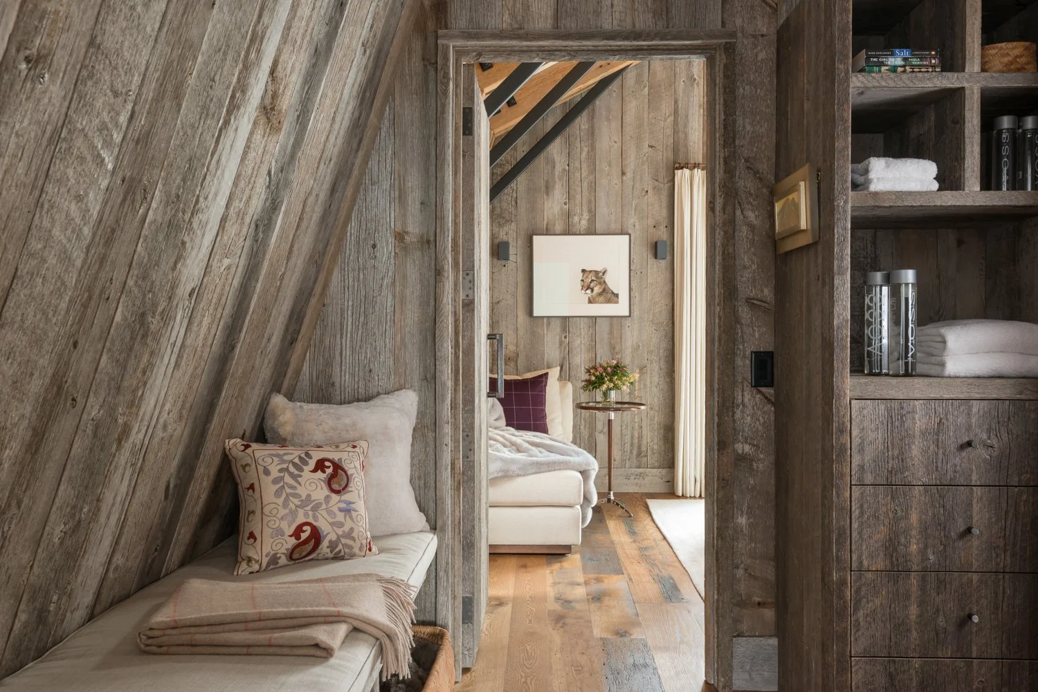 Interior of a rustic bedroom with wooden walls and flooring, featuring a cozy seating area with cushions and blankets, a framed artwork of a lioness on the wall, and a shelf with folded towels and books.