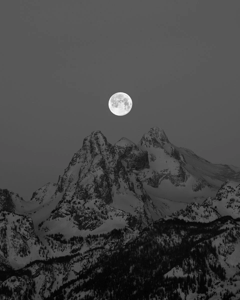 Nighttime mountain landscape with snow-covered peaks and a full moon in the sky.