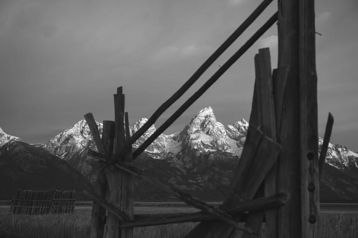 A black and white photo of a broken wooden structure in a field with snow-capped mountains in the background.