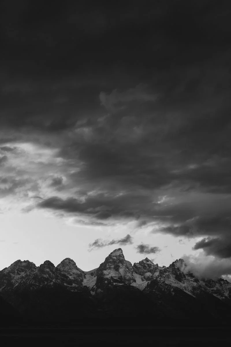 Black and white photo of rugged mountain range with peaks covered in snow and dramatic cloudy sky above.