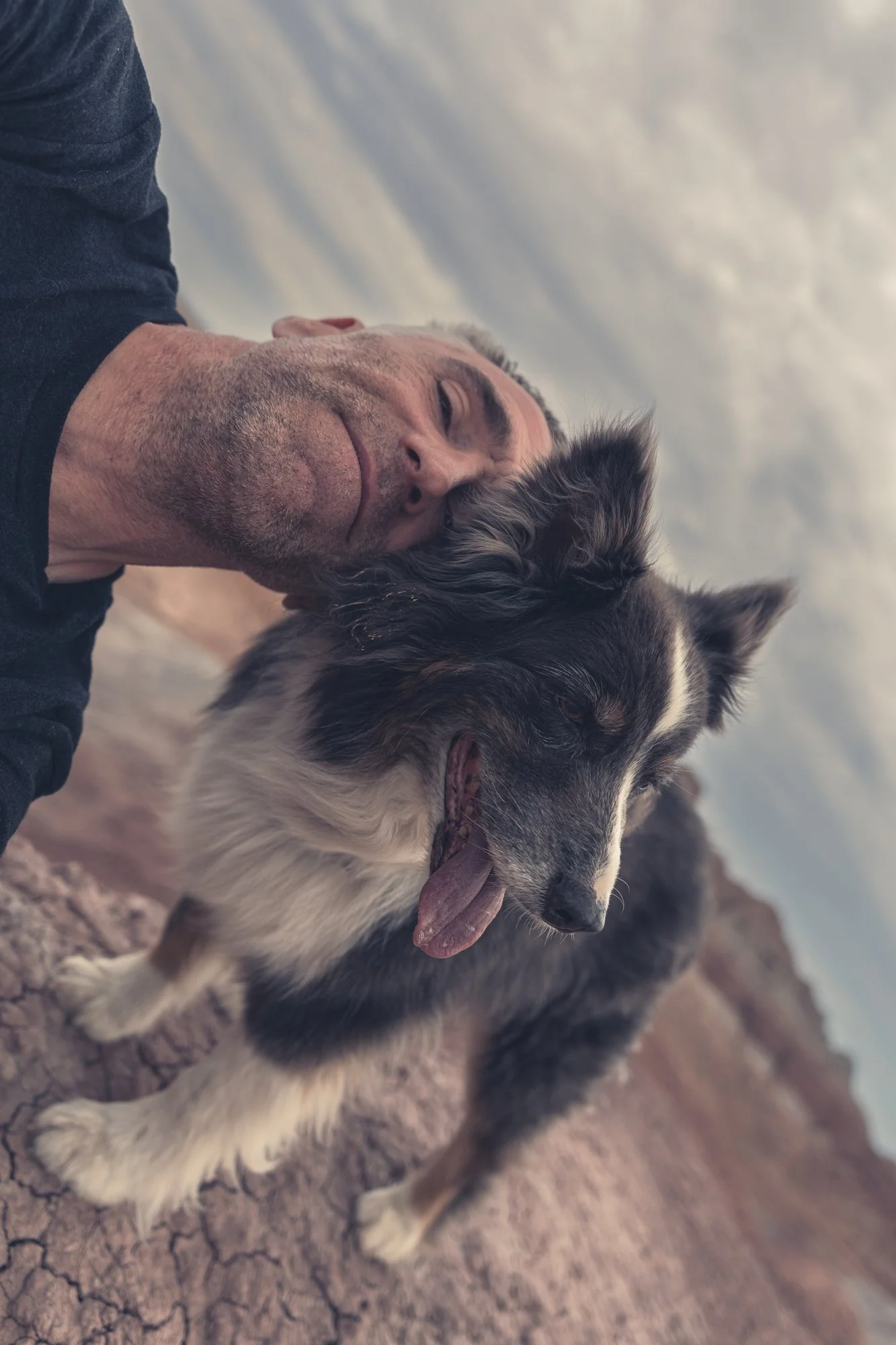 A man taking a selfie with his dog outdoors on a cracked earth landscape during daytime.