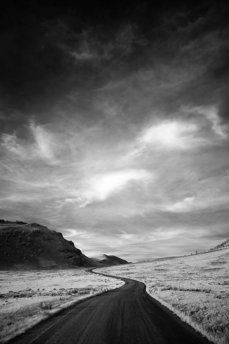 A black and white photo of a winding road in a rural area with hills and grassland on either side, under a cloudy sky.