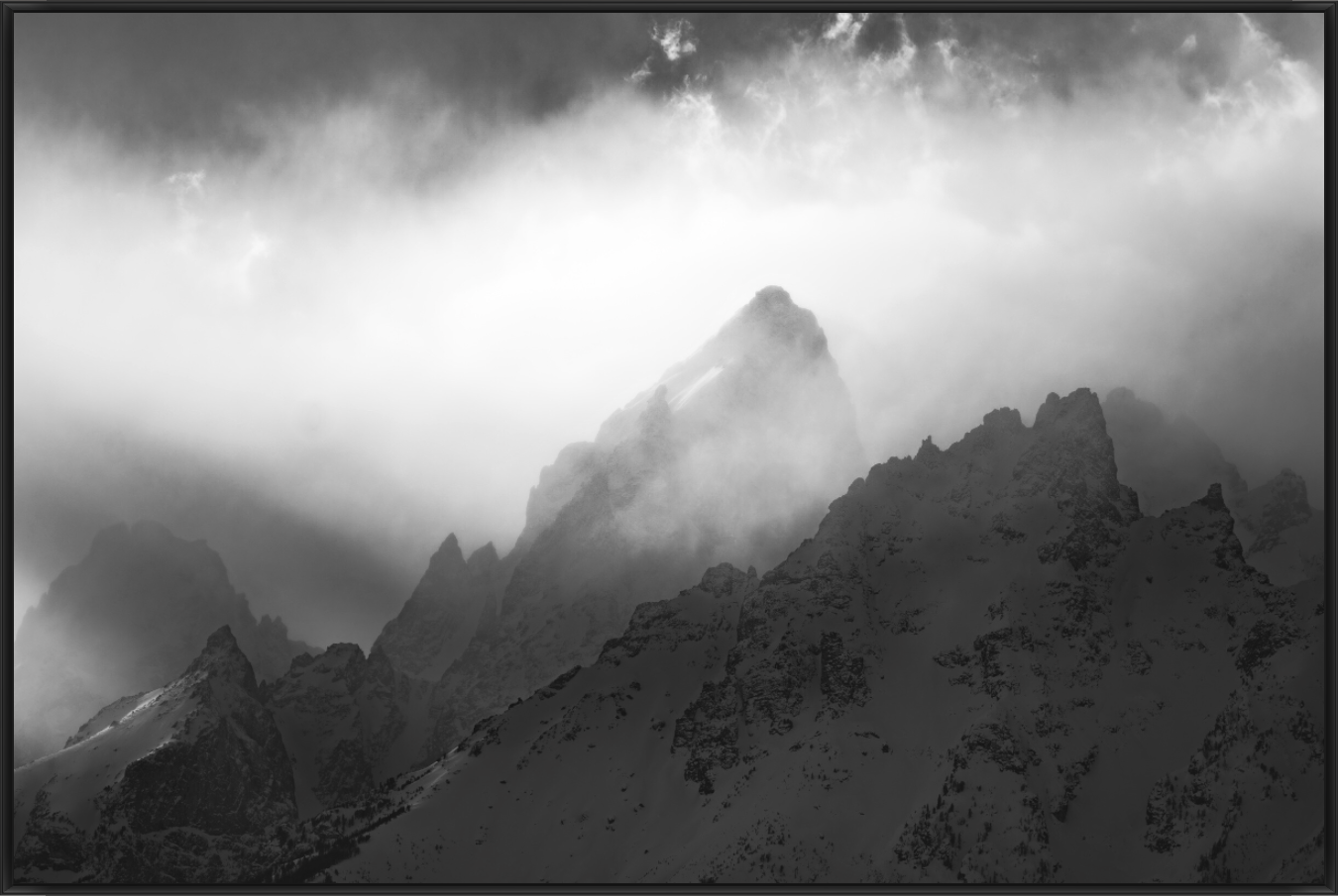 Black and white photo of snow-covered mountain peaks with clouds and mist surrounding them.