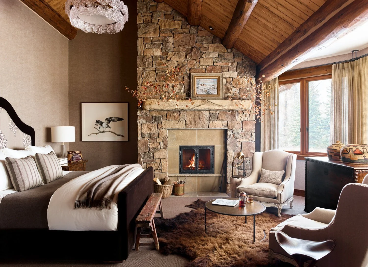Cozy living room with a stone fireplace, beige armchairs, a coffee table, and large windows with curtains, creating a warm and inviting atmosphere.