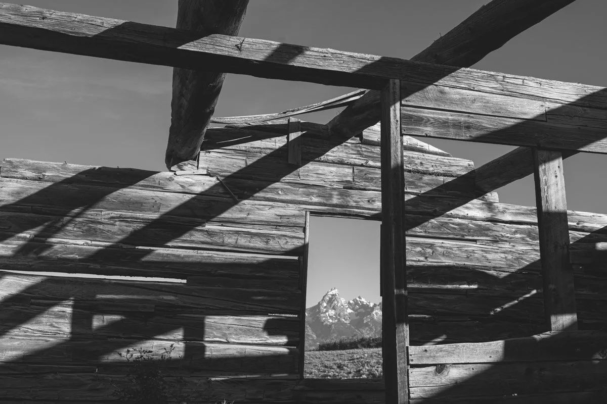 Black and white photo of a partially collapsed wooden structure with a mountain view through an opening in the wall.