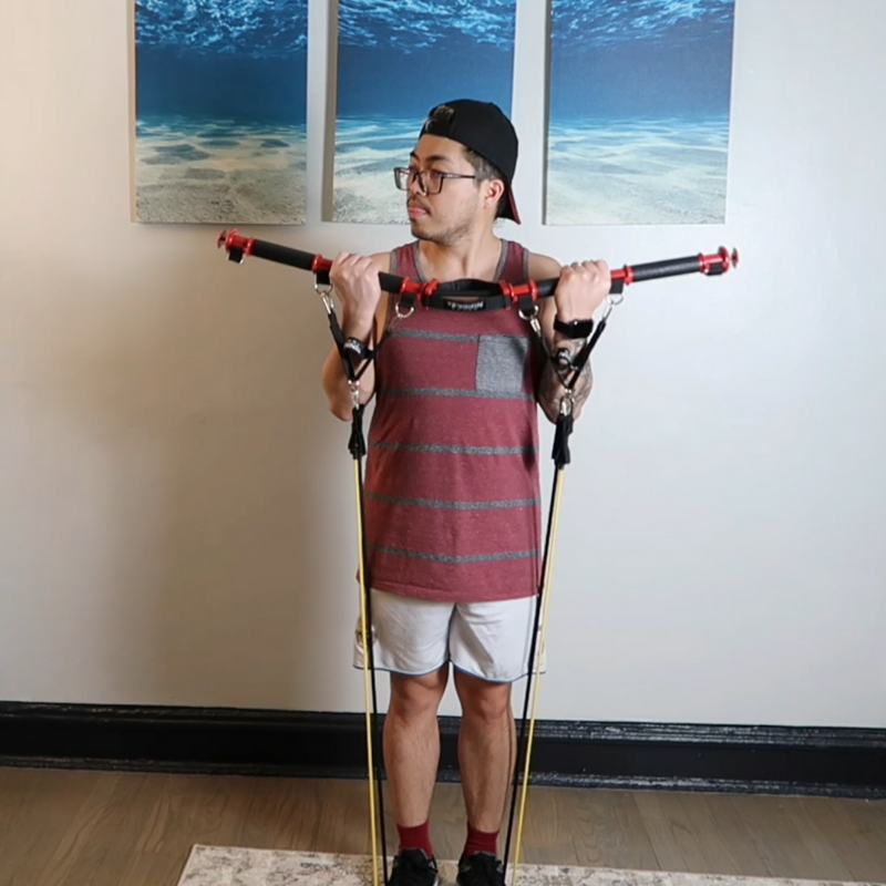 A young man standing indoors, holding a trekking pole across his shoulders, wearing a sleeveless shirt, shorts, a backwards cap, and glasses, with three beach pictures hanging on the wall behind him.