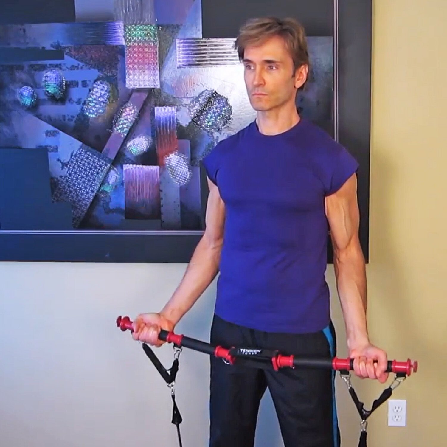 A man holding a red and black exercise bar with straps, standing in front of a colorful abstract wall art in a room with a beige and black wall.