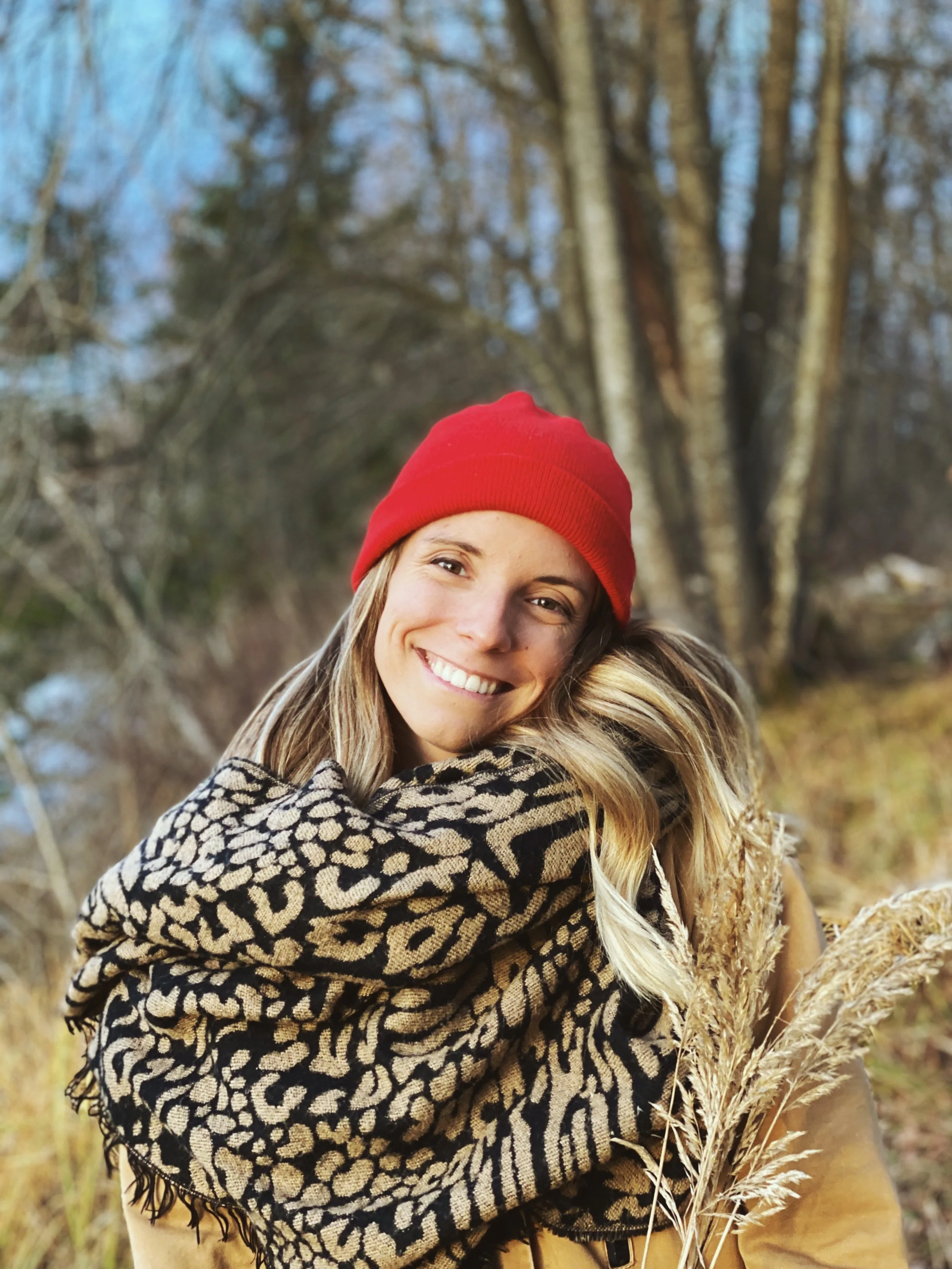 Online prenatal yoga training review. A smiling woman wearing a red beanie and a leopard-print scarf outdoors in a wooded area with tall dry grass.