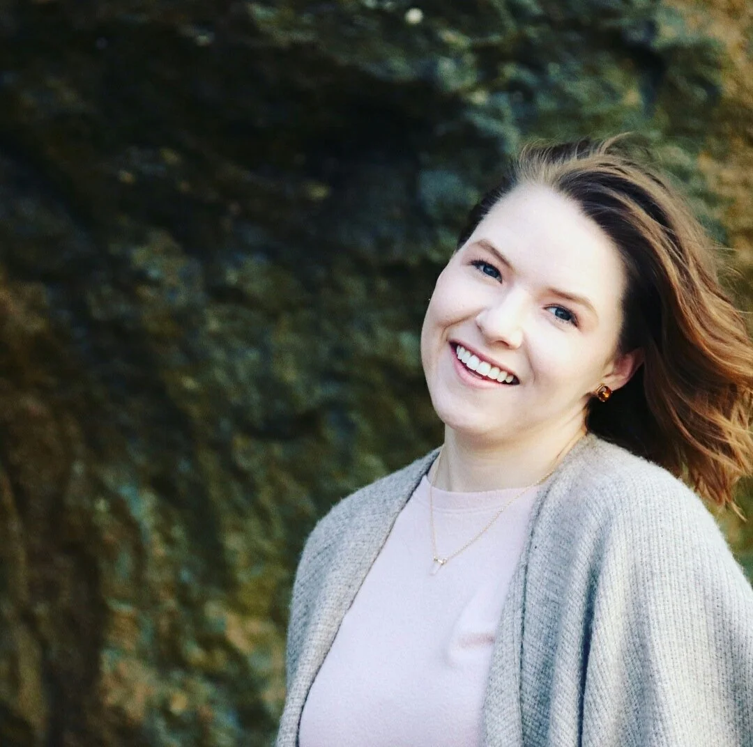 A young woman with light skin and brown hair smiling outdoors with a rocky background.